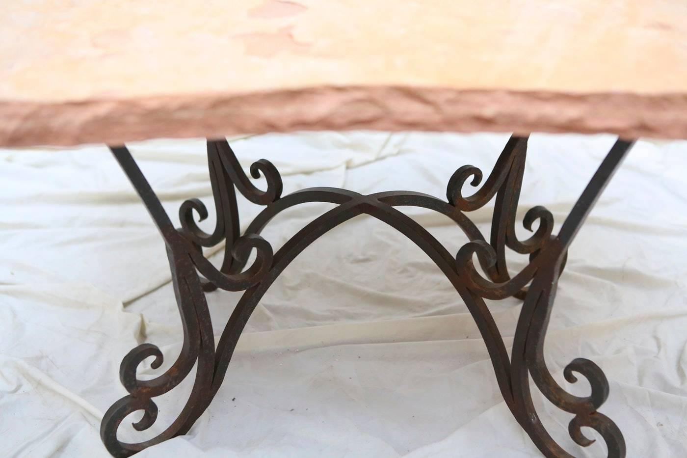 A natural cut stone topped table with a wrought iron base. The tabletop is square with rounded edges cut in. The top sits on a scrolled black wrought iron base.