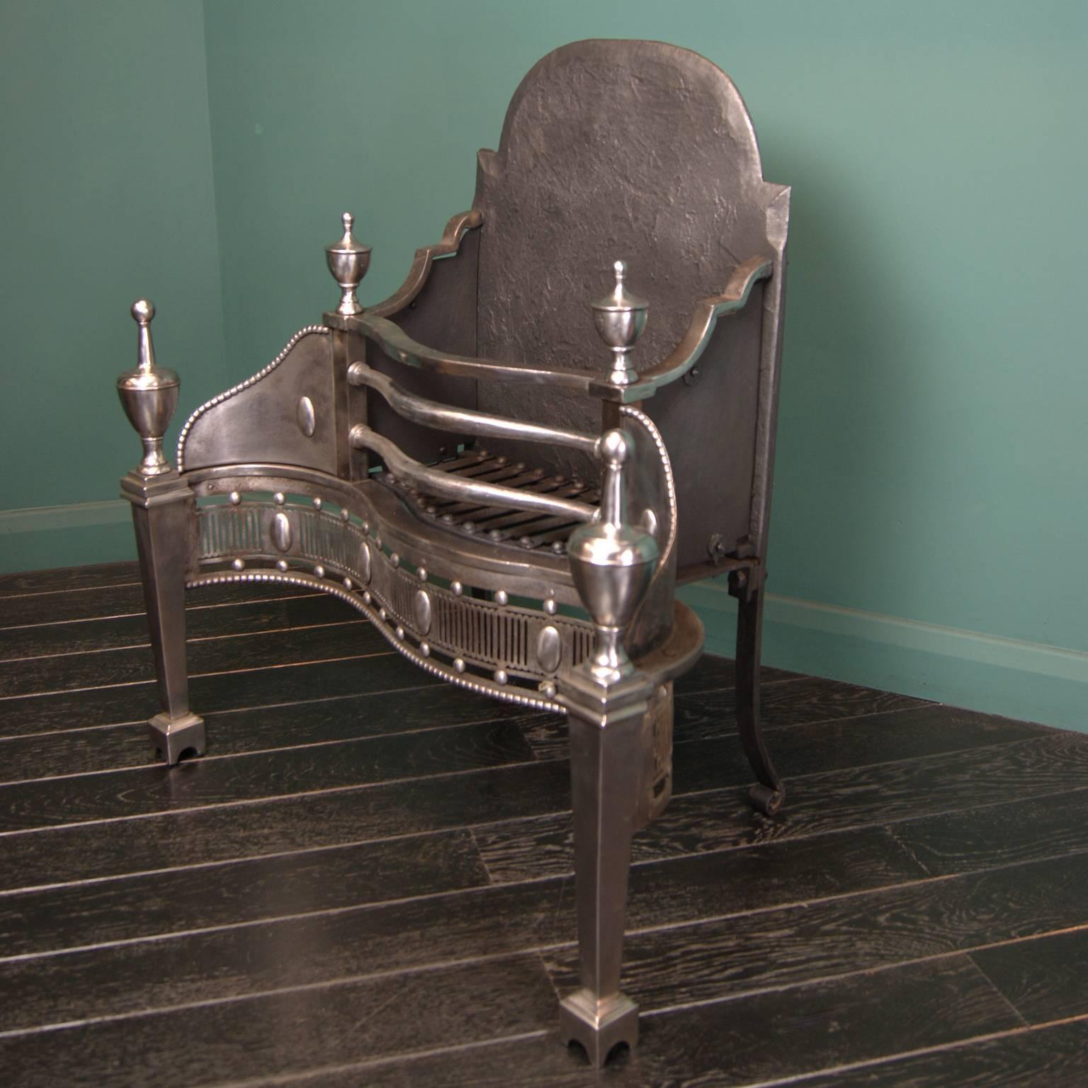 An elegant, tall polished steel dog grate. Tapered legs set on arched plinths under pinacle urn finials. Serpentine front with pierced fluted fret plate interrupted with paterae over beaded moulding. Rear elements consist of rivet-work grill, shaped