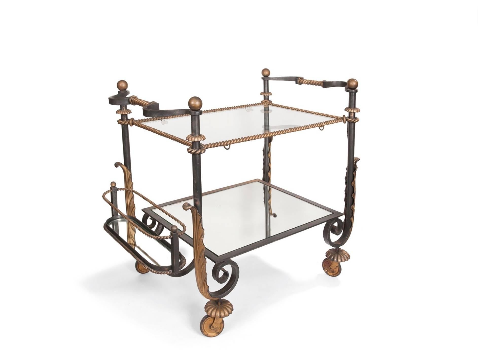 In wrought and gold iron, rectangular shape with two glass plates, the upper plate girded with a golden cord, the plates connected by four tubular uprights ending in spirals outlined by a large gold acanthus leaf, on four wheels topped by a golden