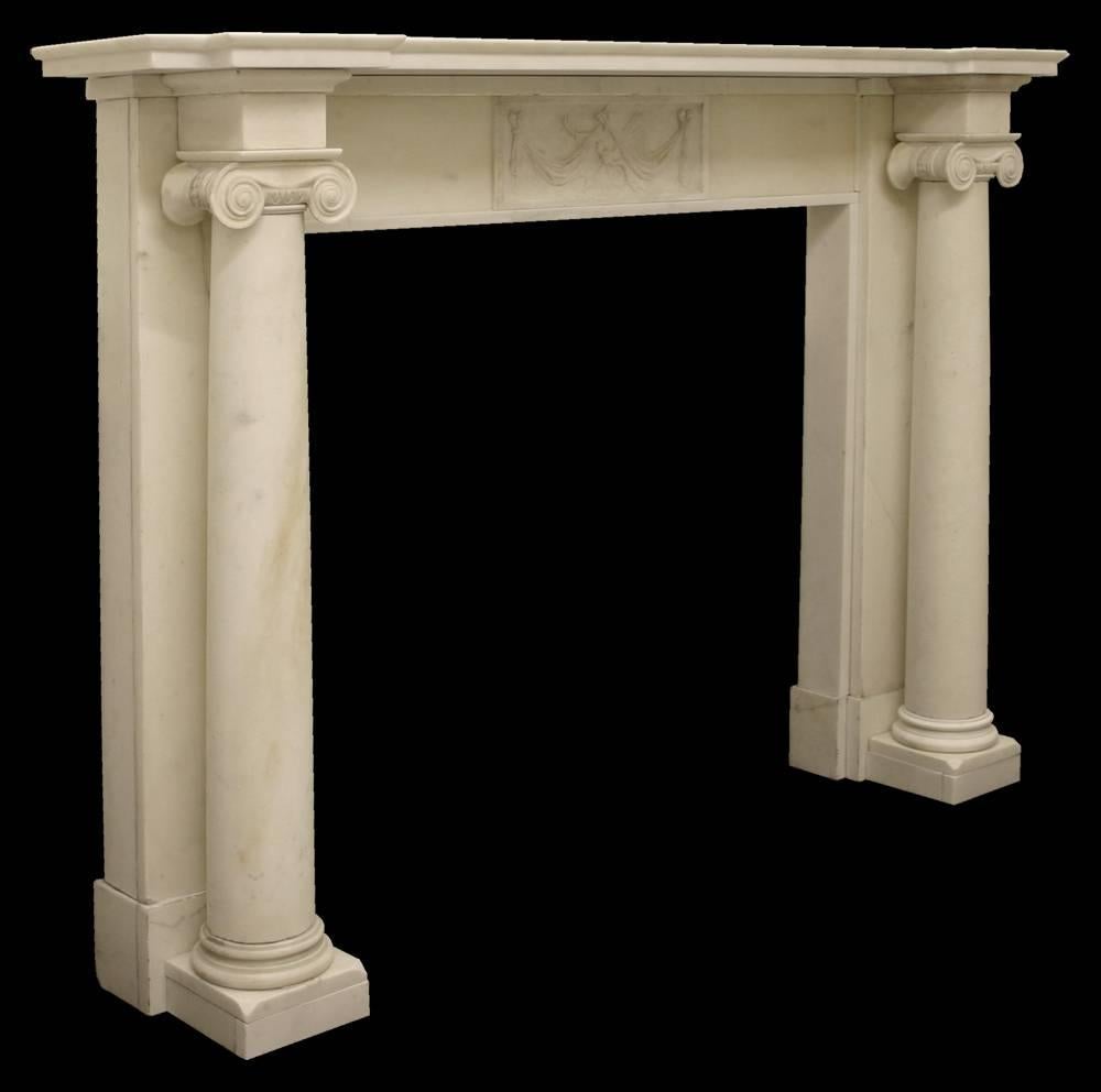 Large and imposing antique Georgian fire surround in statuary marble with full ionic pillars terminating in square plain capitals which in turn support a break front shelf. The frieze benefits from a carved tablet to the centre showing a drape above
