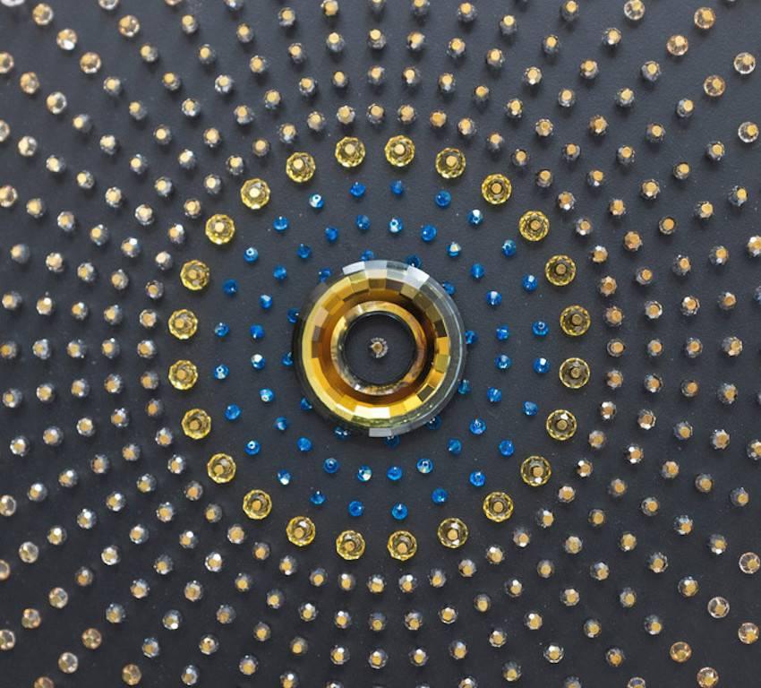 2000 Swarovski crystals are arranged in a intricate pattern around an iridescent, orange and blue crystal ring.
Delicate golden jewellery pieces and indigo blue hand-sown crystals make up elements of this unique mandala-esque sculpture.
The wall