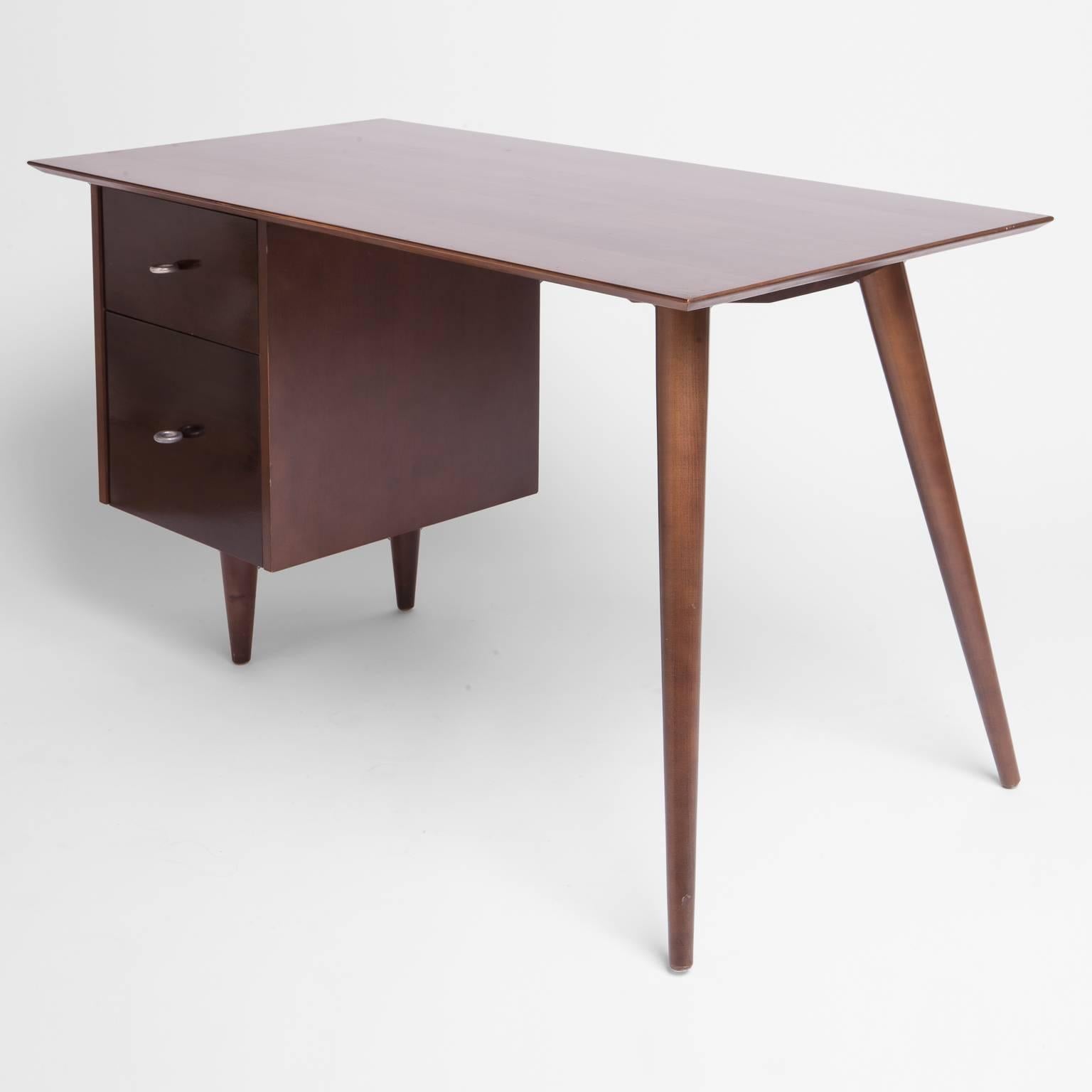 Dark mahogany stained, medium sized desk supported by two drawers to the left and the signature angular tapered legs on the right. There is ample knee space without a center drawer. The top drawer retains the Planner Group label, bottom drawer is