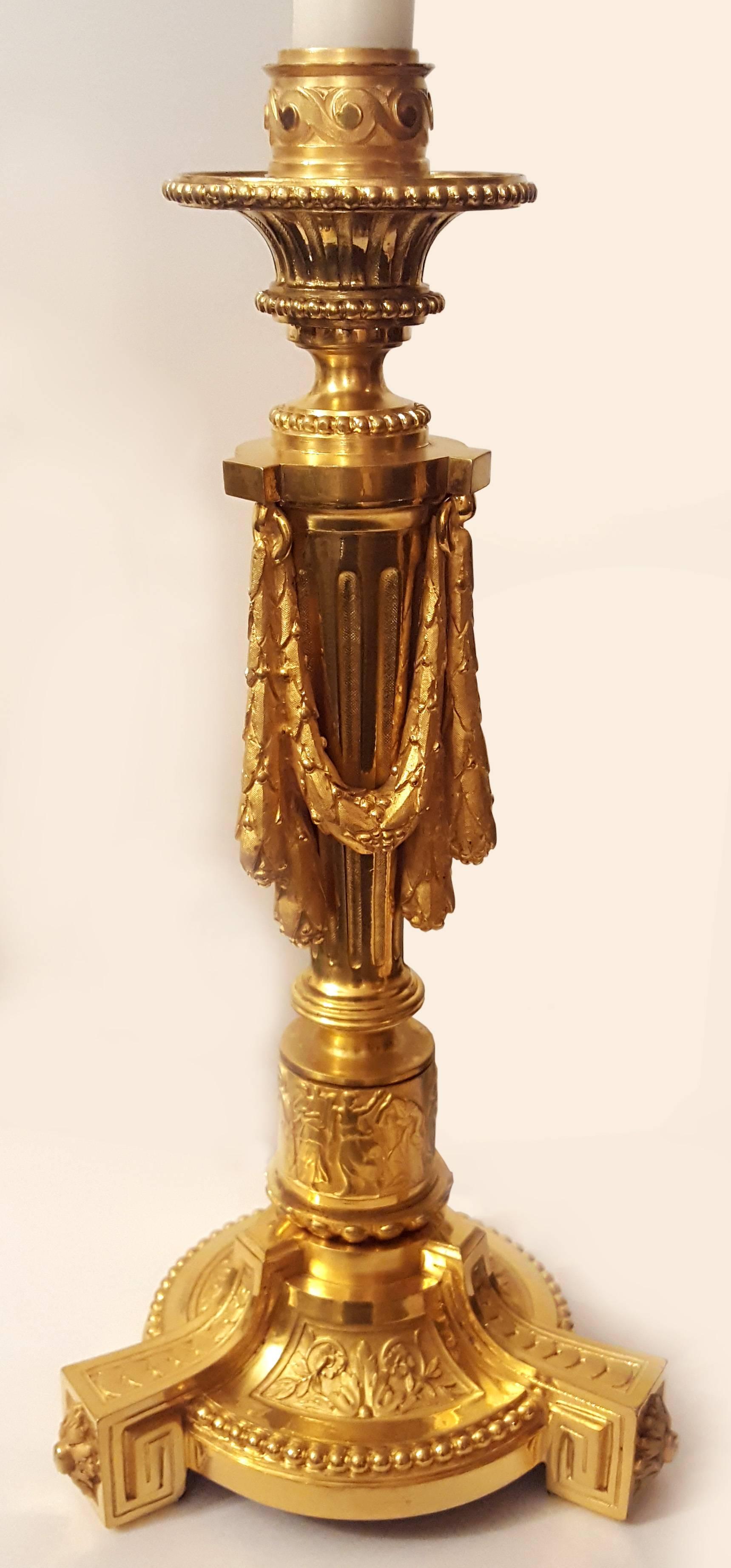 A pair of massive French gilt ormolu tripod candlesticks, mid-19th century.
Adorned with flowers, a Greek column with three garlands, a circumferential relief with vivid figures in ancient Greek dresses, one of them holding a lyre, meanders.