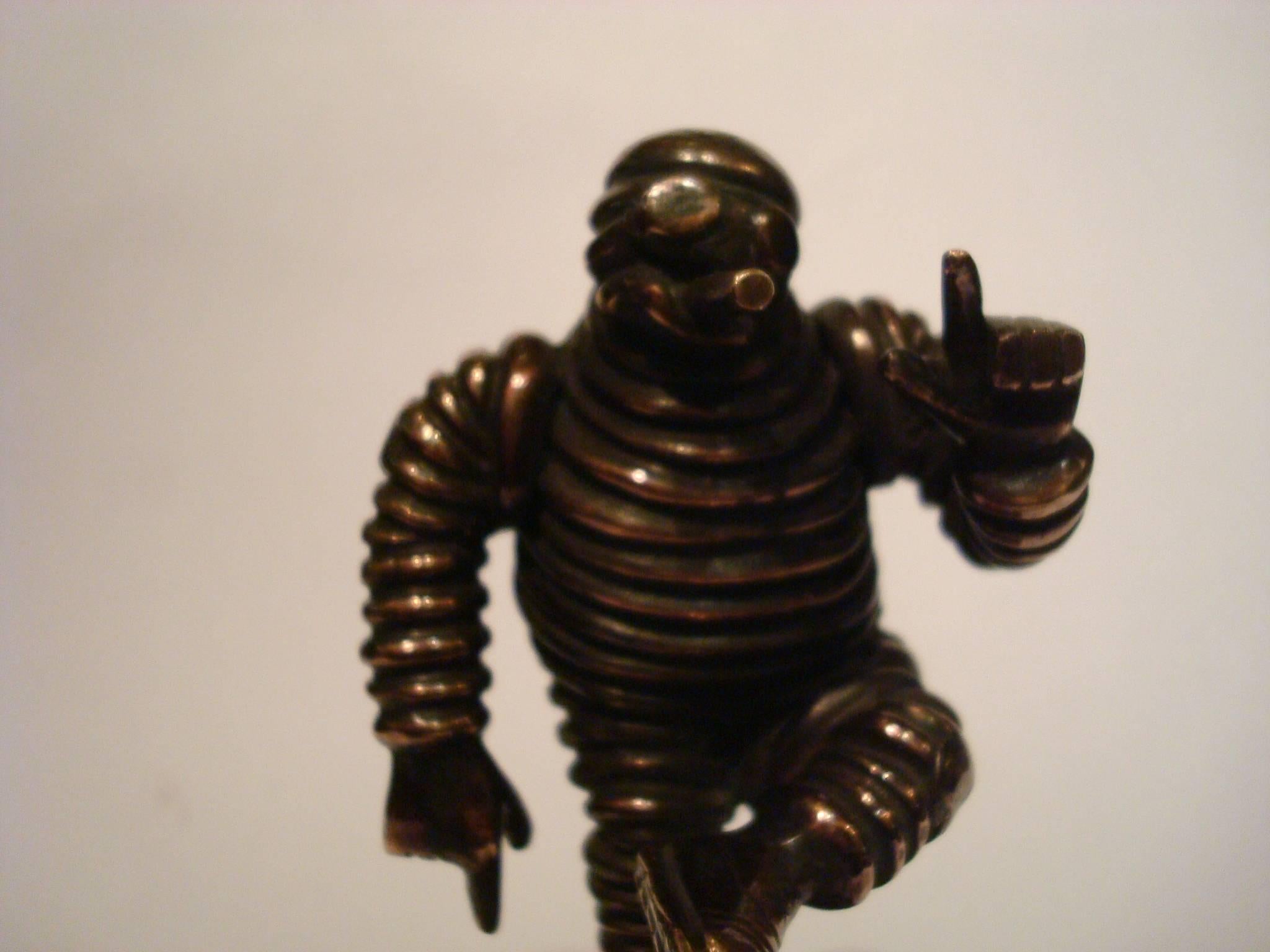 This is a highly collectible Michelin Man bronze car mascot.
It´s approximate 4 inch tall depiction of the Michelin man, also known as Bibendum, joyfully raises his arm while jauntily kicking up his leg. Also recognized as Bib or Bibelobis, the