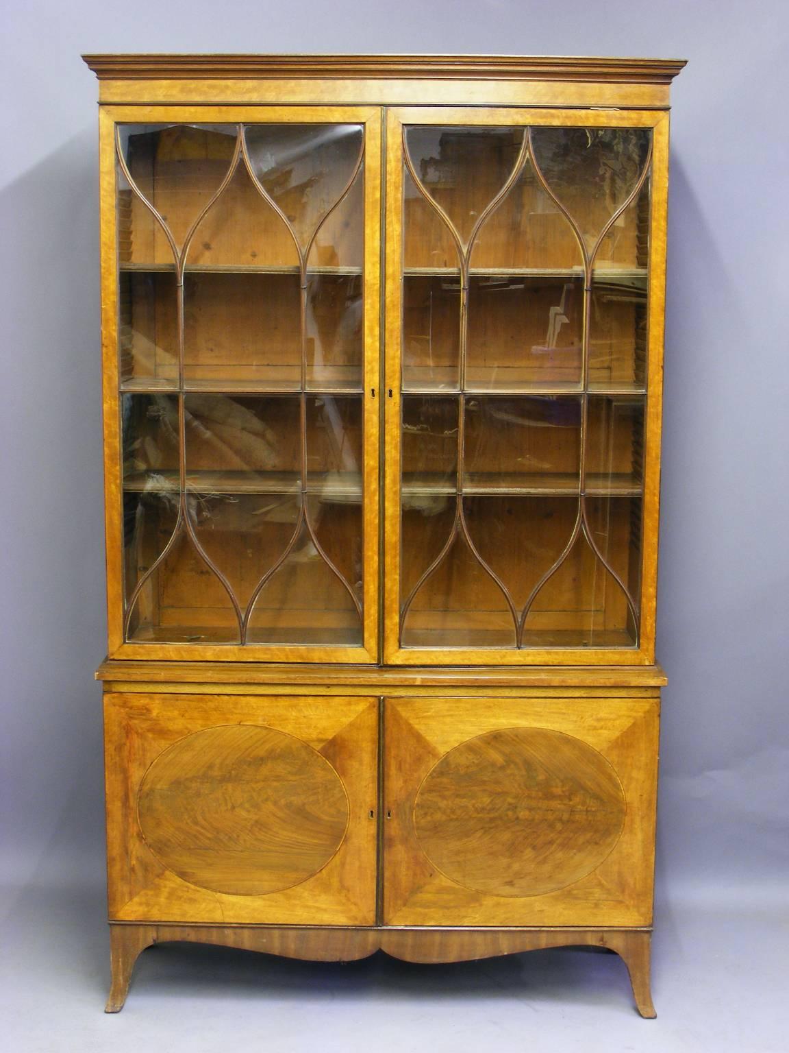 An antique Sheraton satinwood and mahogany display cabinet. The beautifully shaped astragal doors enclose adjustable shelving. The doors of the lower section are inset with flame mahogany panels. The whole is raised upon outswept bracket feet, circa