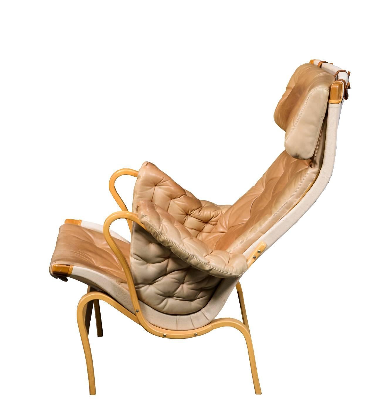 Model Pernilla armchair by designer Bruno Mathsson and produced by DUX. 

The chair has a laminated beechwood frame, stretched with canvas. The chair features its original cushions and armrests upholstered in beige buttoned leather.
   