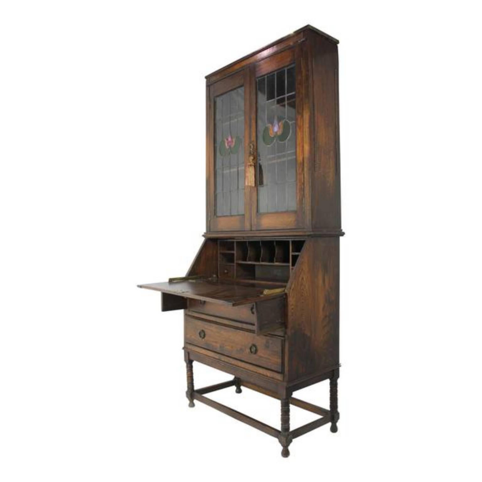 Fold down the writing surface to extend the supports, revealing nine cubby holes and two small drawers and underneath two larger drawers for storage. Lovely arts and crafts cut-glass panels on the upper case with two shelves. Both the desk front and