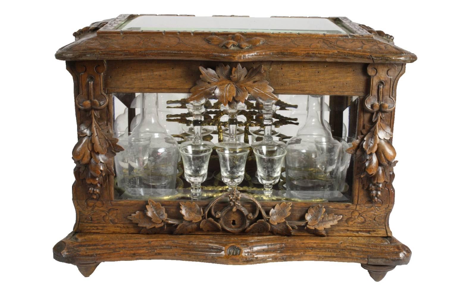 Carved walnut and beveled glass panels in the surround, hold a removable base with four etched decanters and 16 etched cordial glasses held by decorative brass and handle.