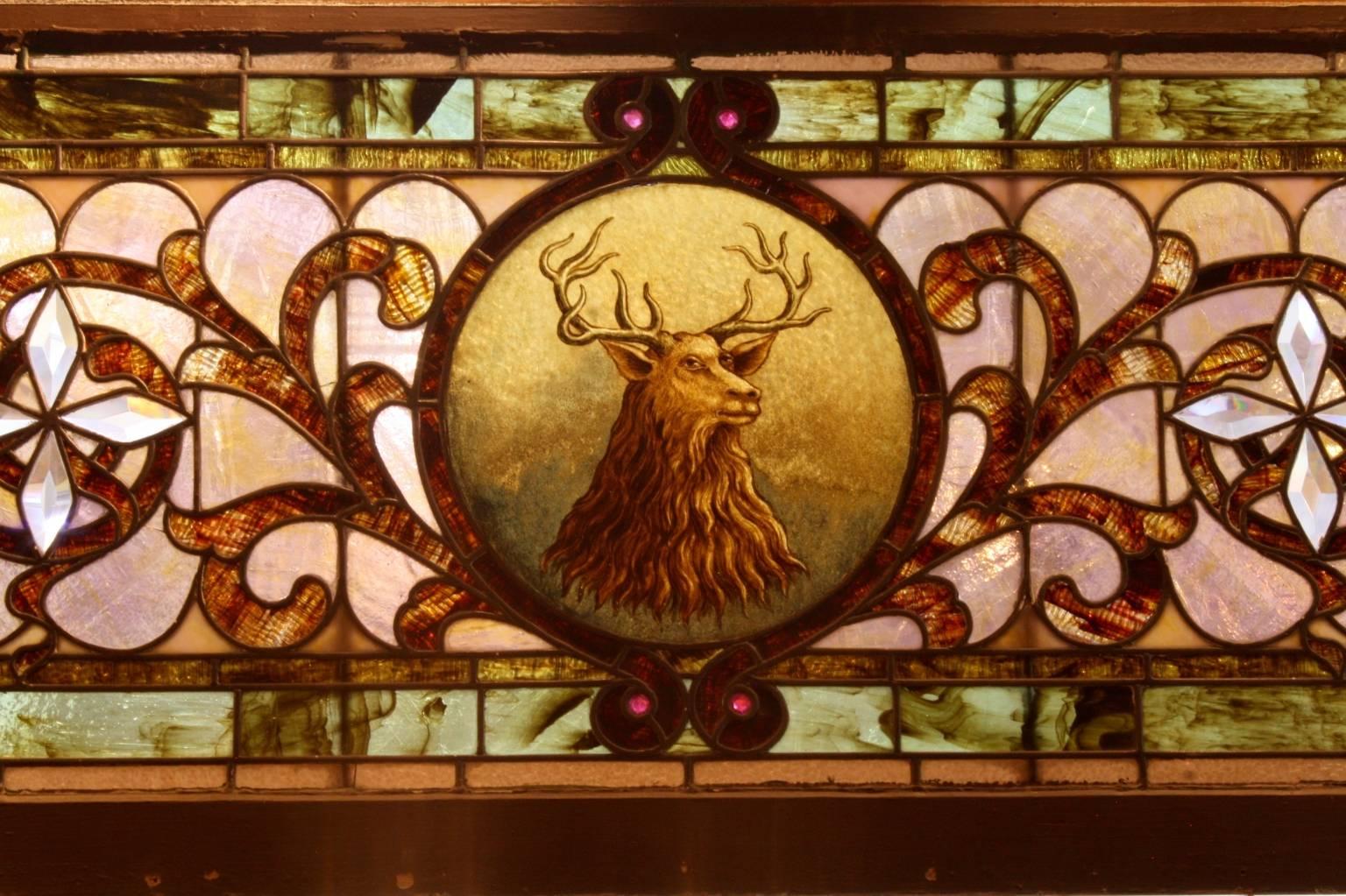 The subtle earth tones of this long German stained glass panel, bright white four pointed stars, vines and beaded ribbons surround the stately, painted red stag with an almost anthropomorphic quality. One of the finest stained glass panels we have