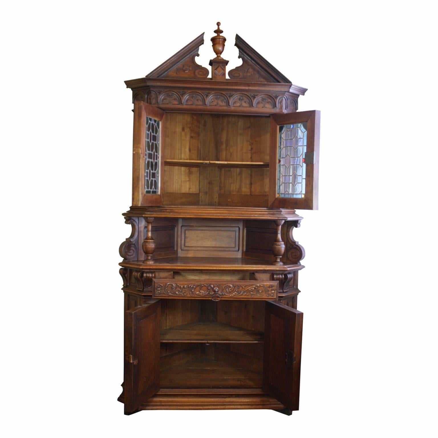 Leaded glass doors accented with blue, red, and gold stained glass are featured on this walnut corner cabinet. The relief carvings of foliage, flowers, and crests, which are masterfully incorporated into the cabinet's design, create a stunning