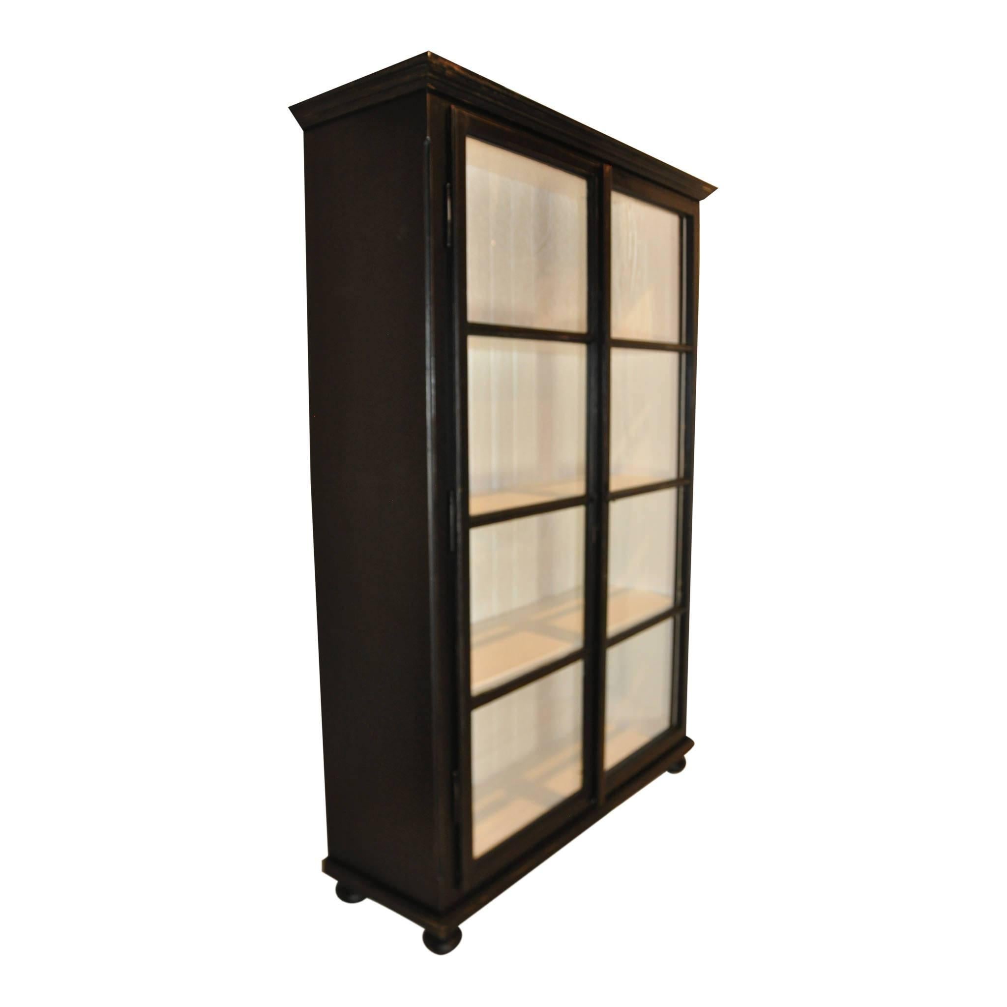 The contrast of the white interior and black exterior of this cabinet makes it a striking piece. Three fixed shelves are visible through the glass panel doors which latch closed. The top is trimmed with crown molding. The cabinet sits on bun feet.
