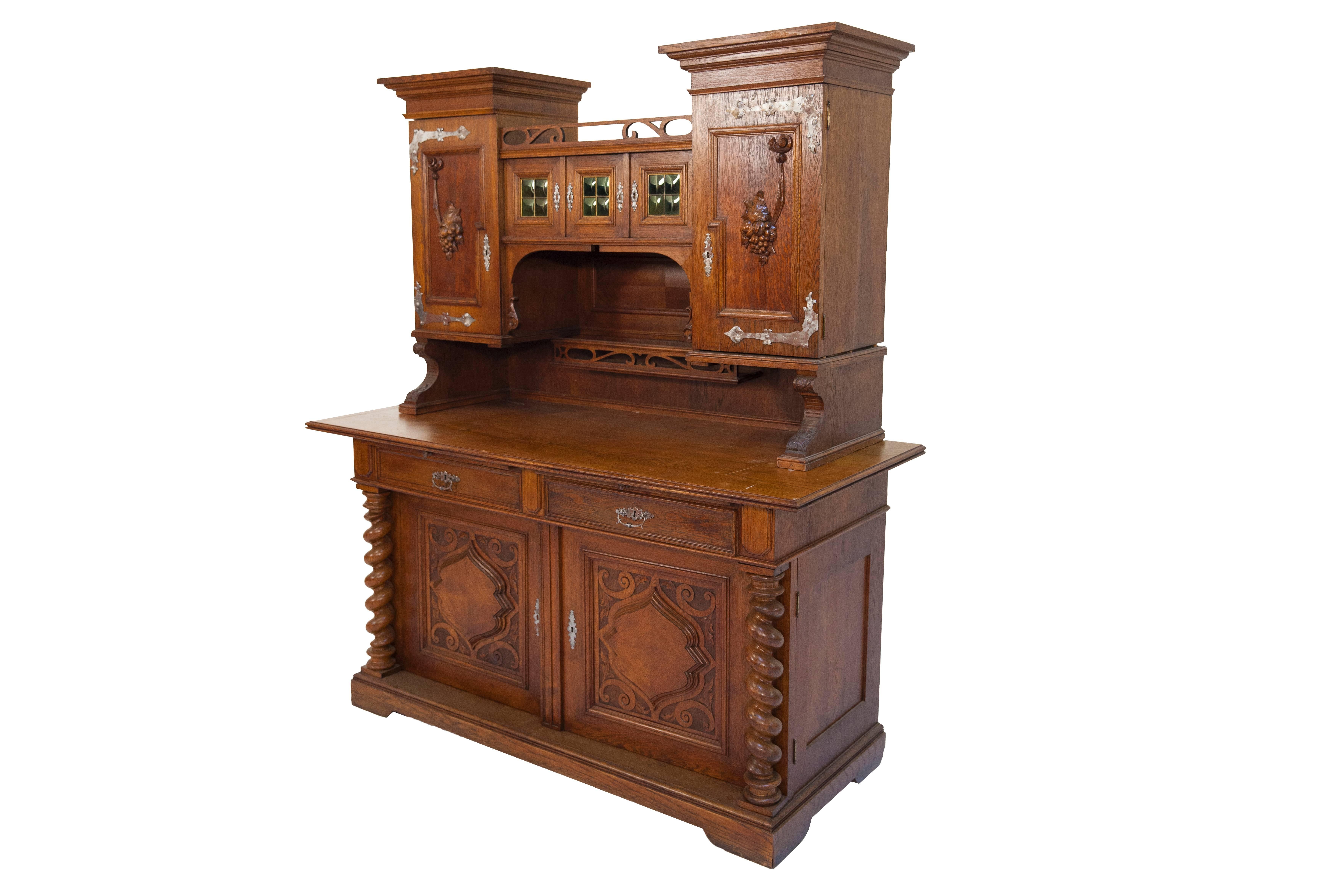 In very good used condition. In its lower part, the sideboard features two doors, with two drawers above; above each of the drawers, a work tray can be pulled out. The panels set into the cut-outs of the doors are doubled-on and framed by carved