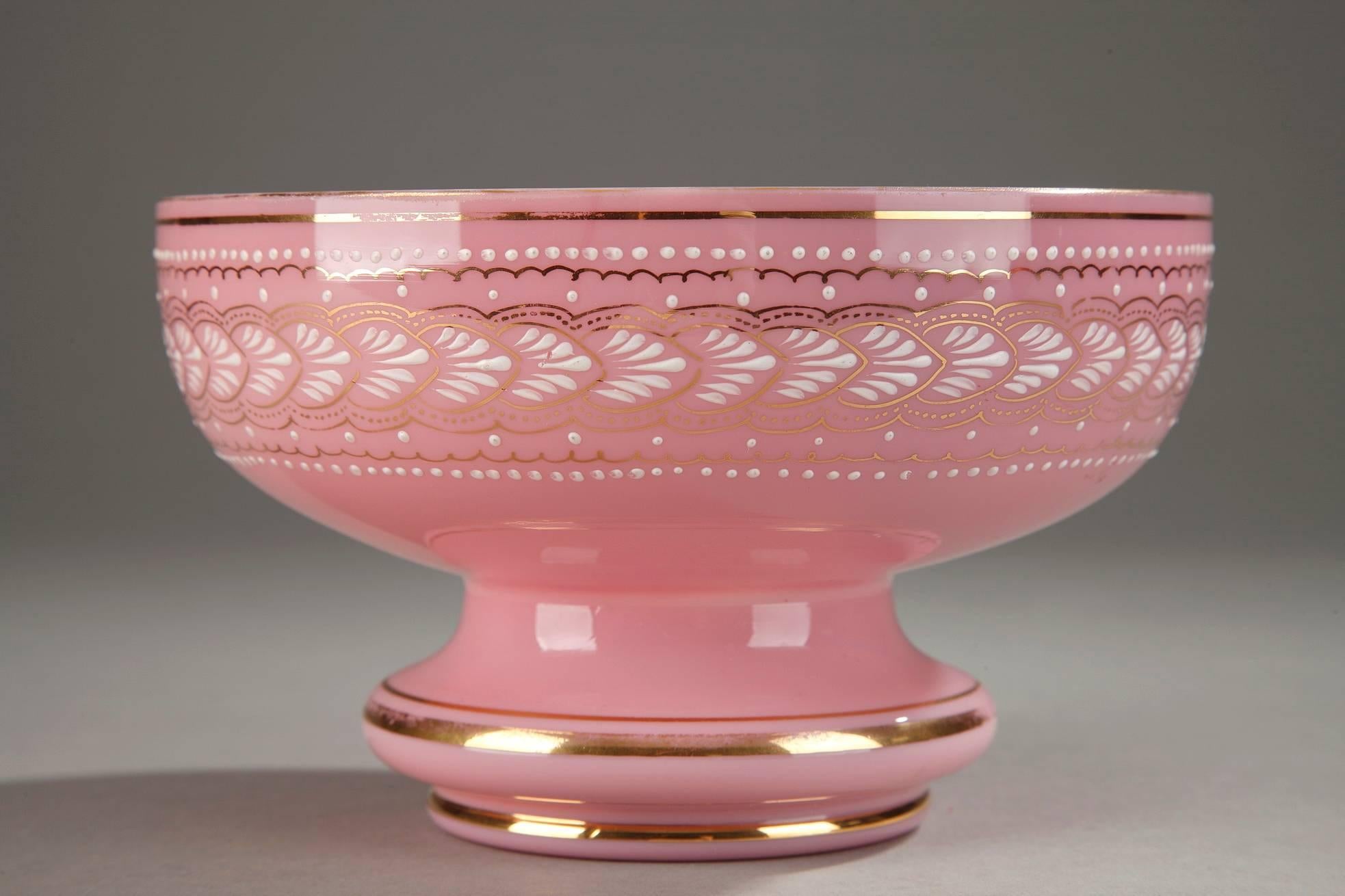 Pink and white opaline bowl decorated on the exterior with a frieze of palmettes, small white dots, and thin, gold embellishments. It rests on a short, pedestal foot that is also accented with gold bands. The white enamel and gold decoration is an