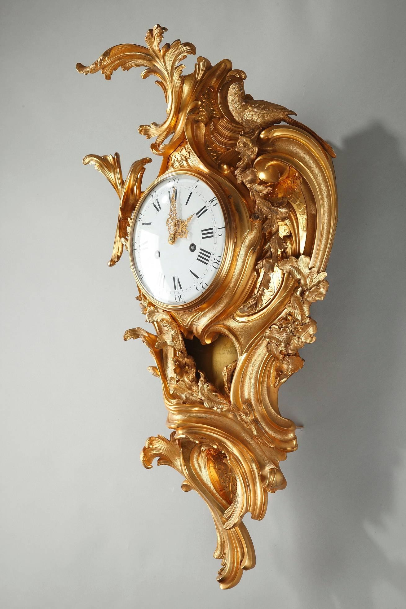 Large, Louis XV style wall clock in gilt bronze. It is richly decorated with foliage and cascades of flowers that are sculpted with deep lines. A sculpted bird sits atop the bountiful ornamentation of this piece. The enameled, white clock face marks