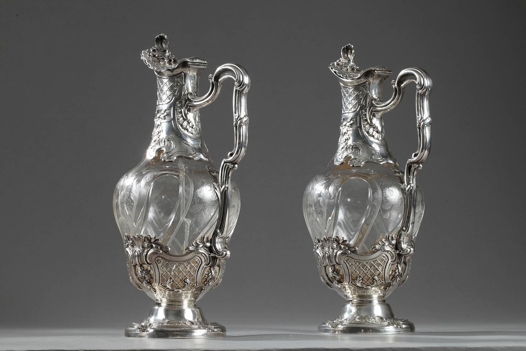 Pair of ewers made of cut crystal and set in silver mounts. The crystal body is engraved with wide curving grooves, flowers, and foliated branches. The silver neck and the base are decorated with sculpted latticework, shells, and foliage. The lid is