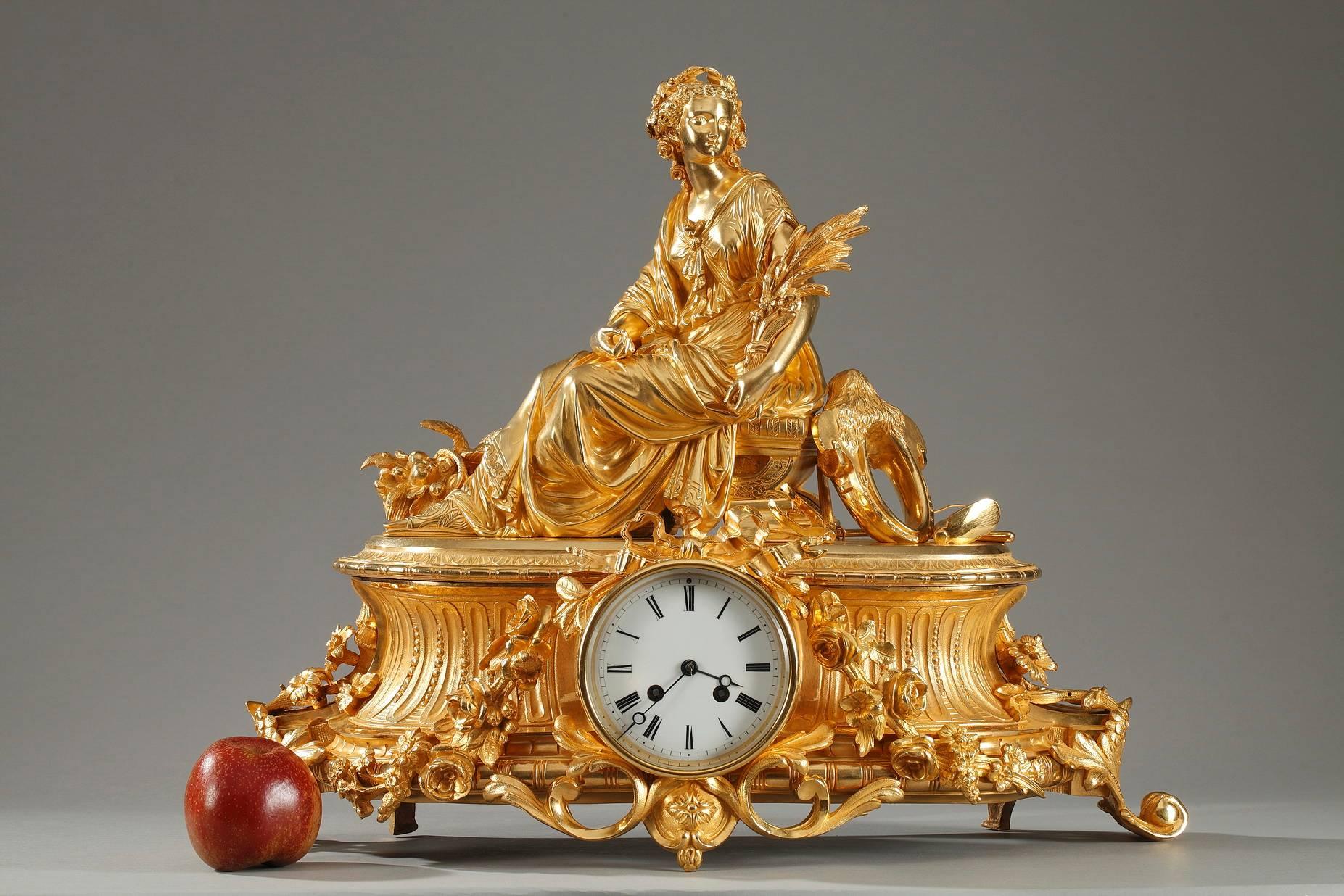 Large, gilt bronze mantel clock in Louis XVI style. The clock is decorated with the figure of Ceres, the Roman goddess of agriculture and fertility. She is resting on a curule seat and is surrounded by a basket of fruits and harvest tools. The oval
