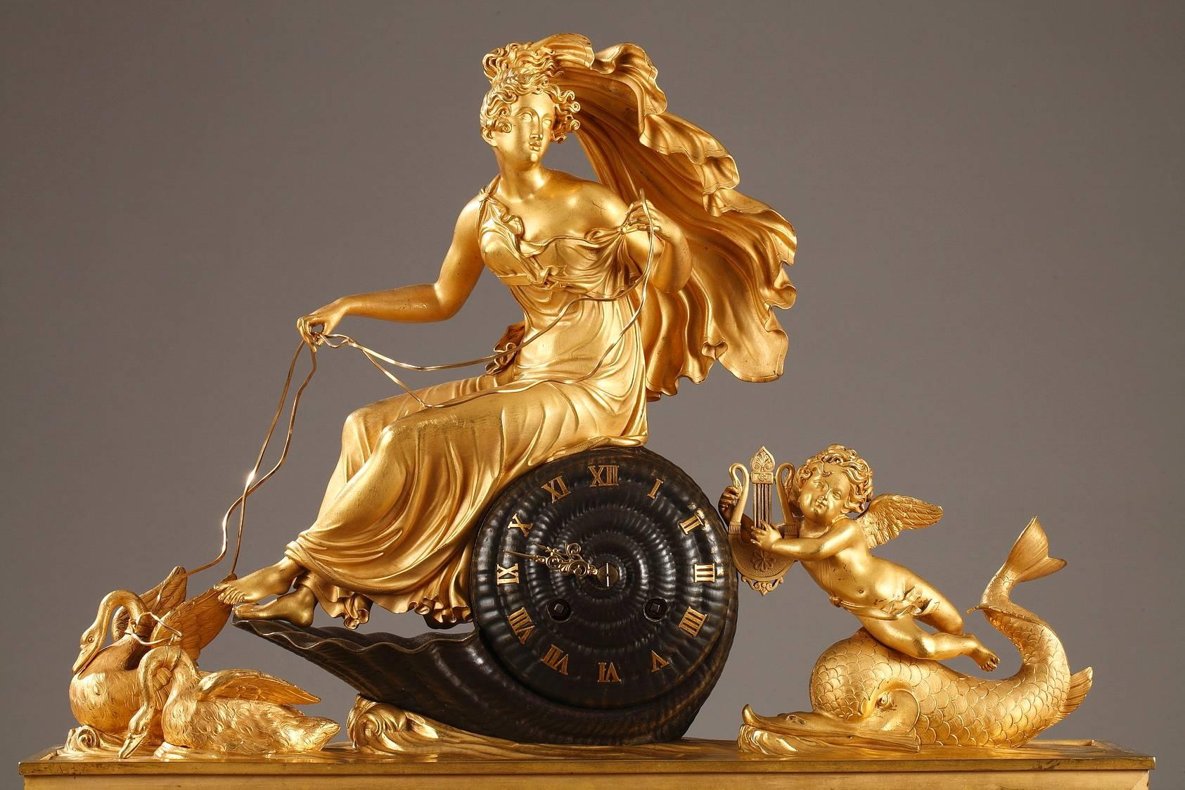 Early 19th century gilt and patinated bronze mantel clock. A young woman is seated on a shell that displays the clock dial, with Roman numerals for hours. Two swans pull the shell and Cupid on a dolphin push it. The set rests on a rectangular base