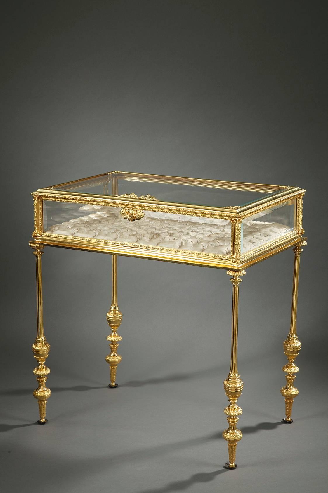 Gilt bronze and glass Napoleon III display case in neoclassical taste. It is decorated with friezes of laurel leaves, small flowers, and angels holding garlands. The case is supported by four slender, fluted legs that descend to meet three foliated