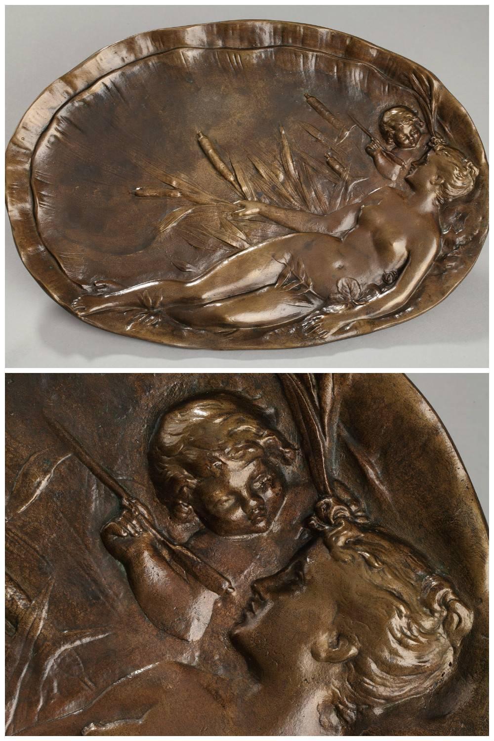 Oval platter in patinated bronze decorated with a sculpted, bas-relief woman stretched out among reeds and cattails. She is nude, and behind her, a young cupid is gazing at her face her through the reeds. This scene, saturated with the sensuality of