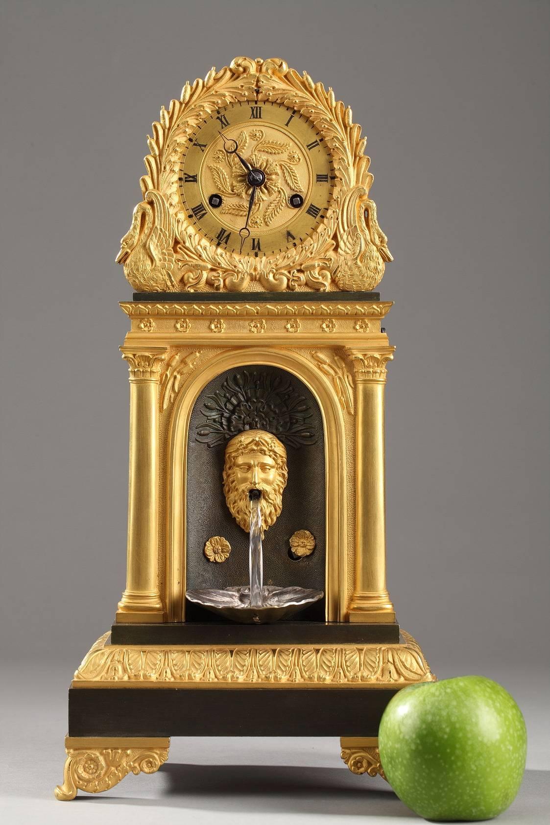 Gilt and patinated bronze mantel clock with a faux fountain. A niche under the clock face houses the face of a river god and a shell. A bar of crystal connects the open mouth of the face to the shell, creating the appearance of the flowing water of