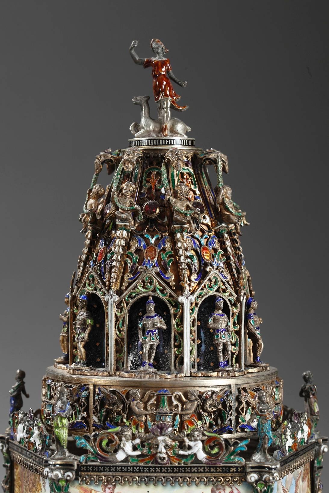 Viennese enamel and silver clock table clock in multicolored enamel and silver. The base is decorated with mythological scenes that represent Diana’s romances and the rape of Europa by Zeus. The scenes are separated by silver pilasters that are