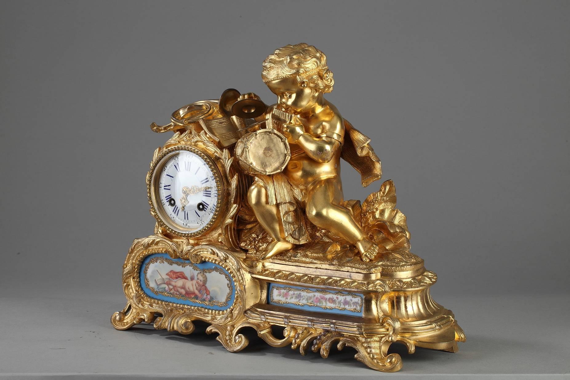 Gilt and sculpted bronze mantel clock featuring a young cupid seated on a rock playing various musical instruments. He is set on a lively base embellished with scrollwork and foliage. The main face of the base is decorated with two