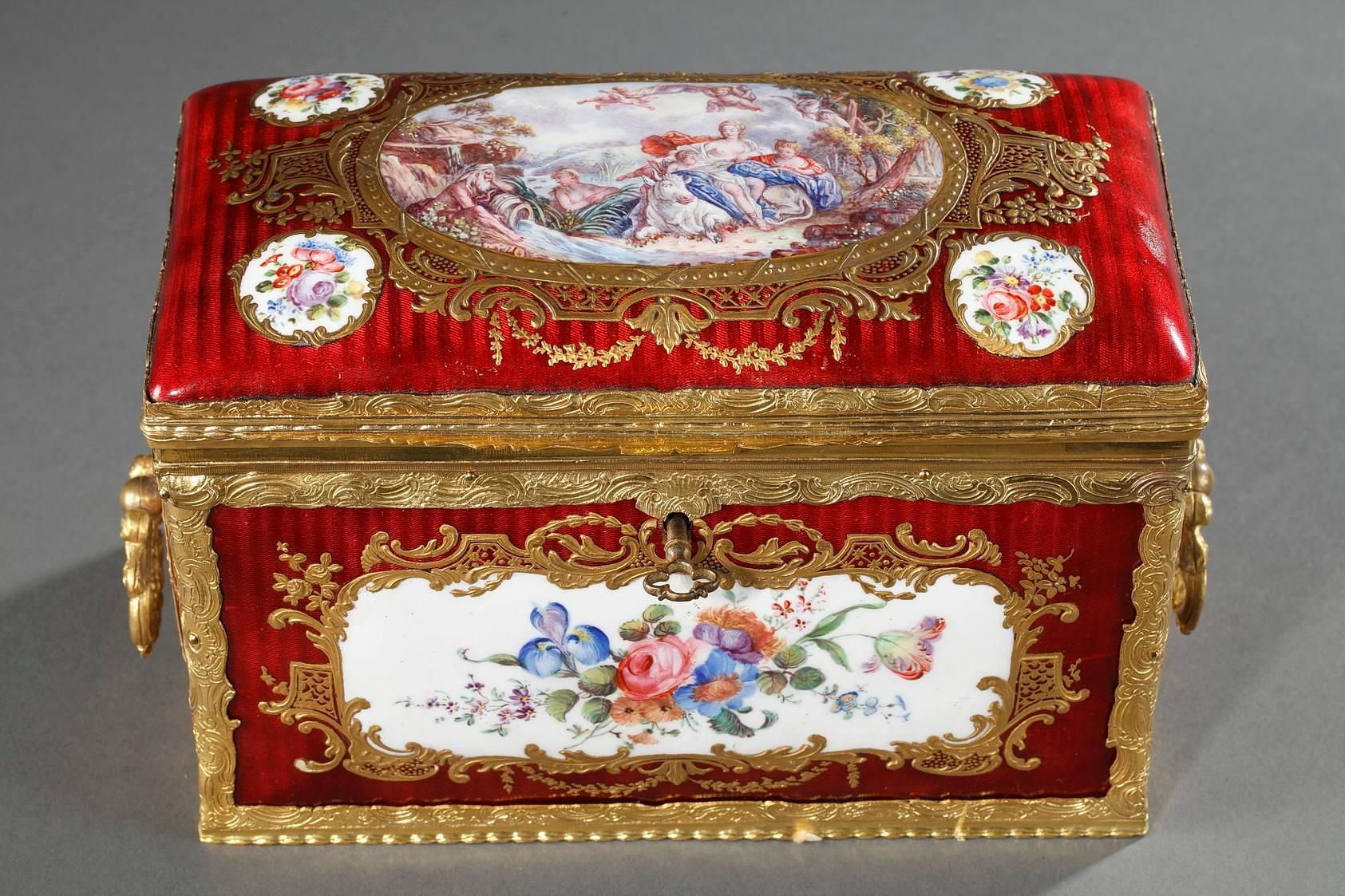Rectangular, red enamel keepsake box with a gilt bronze frame that is very elaborately sculpted with leafy scrollwork. The body of the box is decorated with medallions featuring multicolored bouquets of flowers on a white background. The medallions