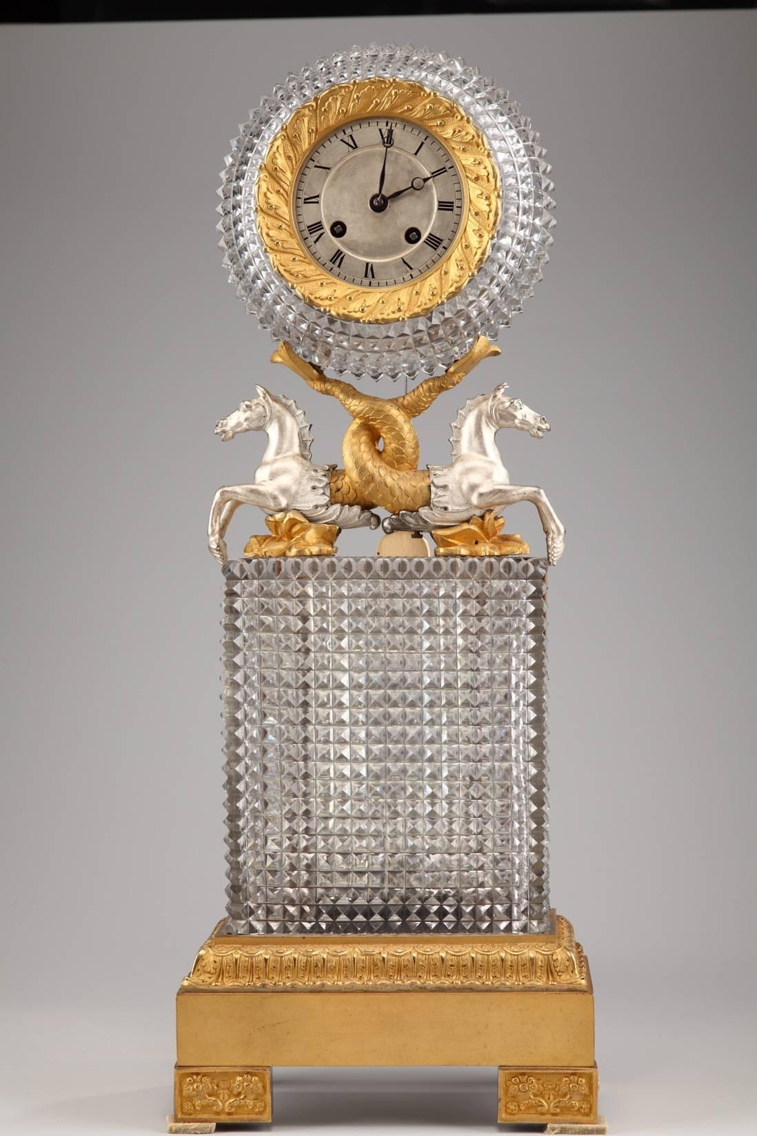 Sizeable Charles X mantel clock composed of a high, cut-crystal base topped by two silvery bronze horses whose torsos transform into intertwining, gilt bronze tails. The horses support the silvery bronze dial, with Roman numerals marking the hours.