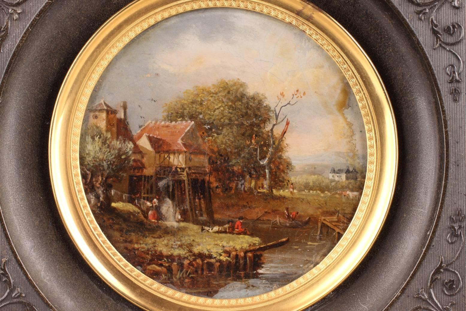 Round, reverse glass painting with its frame, featuring a countryside scene near a river. In the foreground, two young fishermen sit on the grass. Behind them to the left, a man and a woman are working in front of a mill that is surrounded by trees.