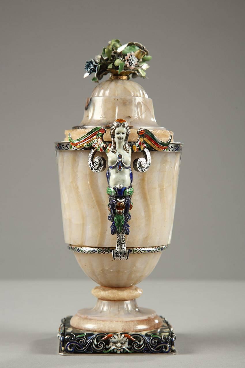 Agate vase decorated with long, fluted canals, gadroons, and an enamel frame adorned with interlacing and flowering scrollwork. The vase is crowned with multicolored flowers and rests on a square base embellished with masks and foliated rinceau. Two