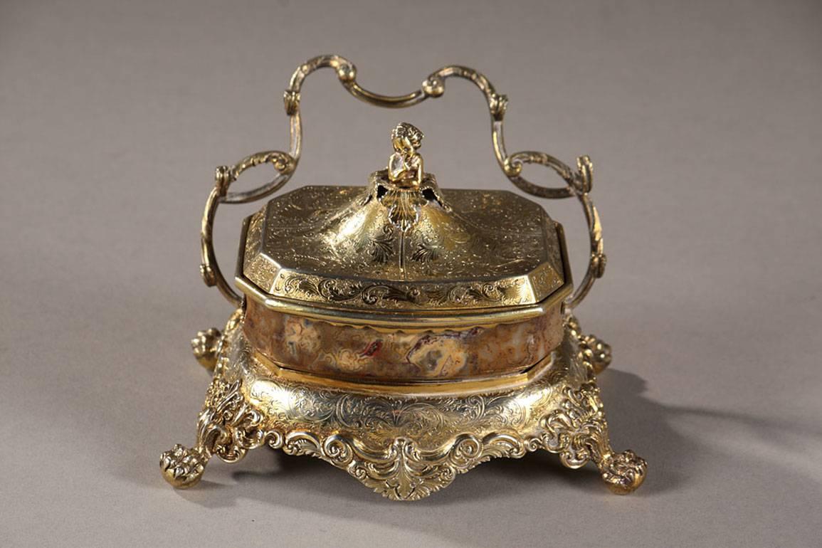 Inkstand in silver-gilt and agate, resting on a flared base. The base is intricately sculpted with foliated rinceau, scrollwork, and acanthus leaves and is supported by four lion paws on balls. The rear feet support the handle, which is decorated