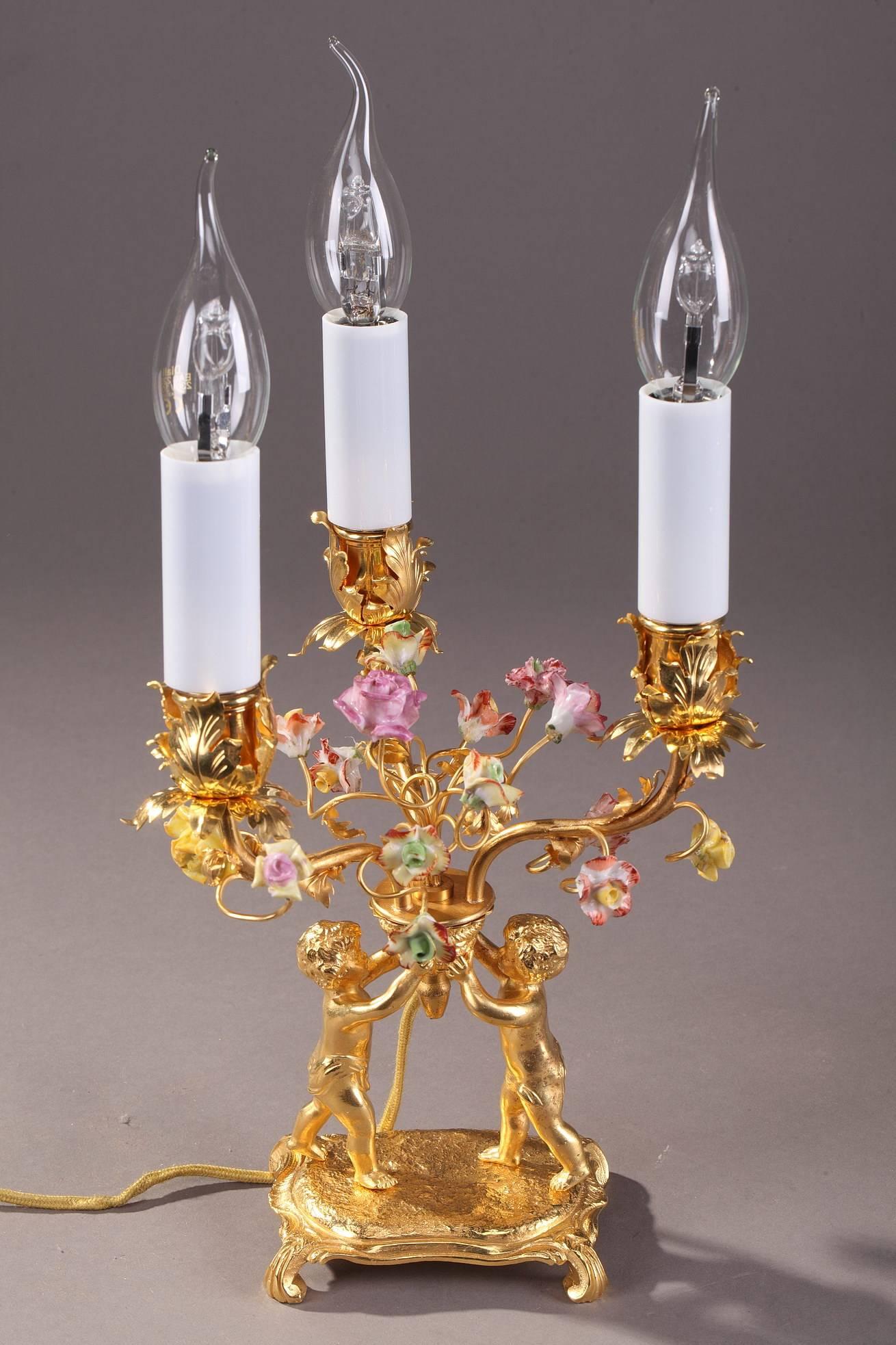 Pair of gilt and chiseled bronze candelabras with three branches each. They are decorated with two putti holding a basket with three candle branches highlighted with porcelain flowers and gilt bronze foliage. Each putto rests on a square, Rocaille