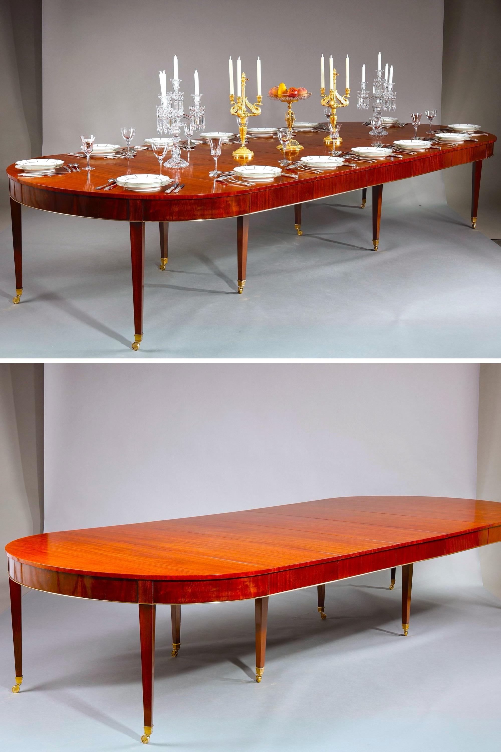 Dining table in solid mahogany, with elegant rounded belt and removable top opening out into five independent extensions. The ensemble rests on eight tapered legs terminating in gilded bronze sabots on casters, facilitating movement and ensuring