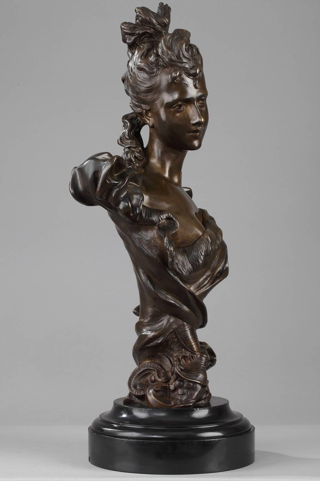 Bust of a woman in nuanced, patinated bronze attributed to the Belgian sculptor Georges Van der Straeten. She is wearing an elegant dress with a ribboned neckline, flowing capped sleeves, and a sash that crosses at her waist. Her lightly curly hair