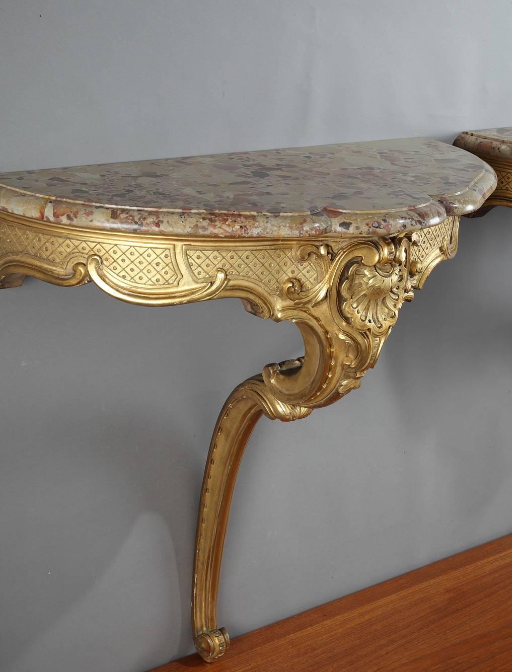 Pair of rounded console tables in giltwood in Louis XV style. They feature sculpted palmettes, foliated scrollwork and a latticework pattern. They are topped with Breche marble from Alep. The two tables are numbered 