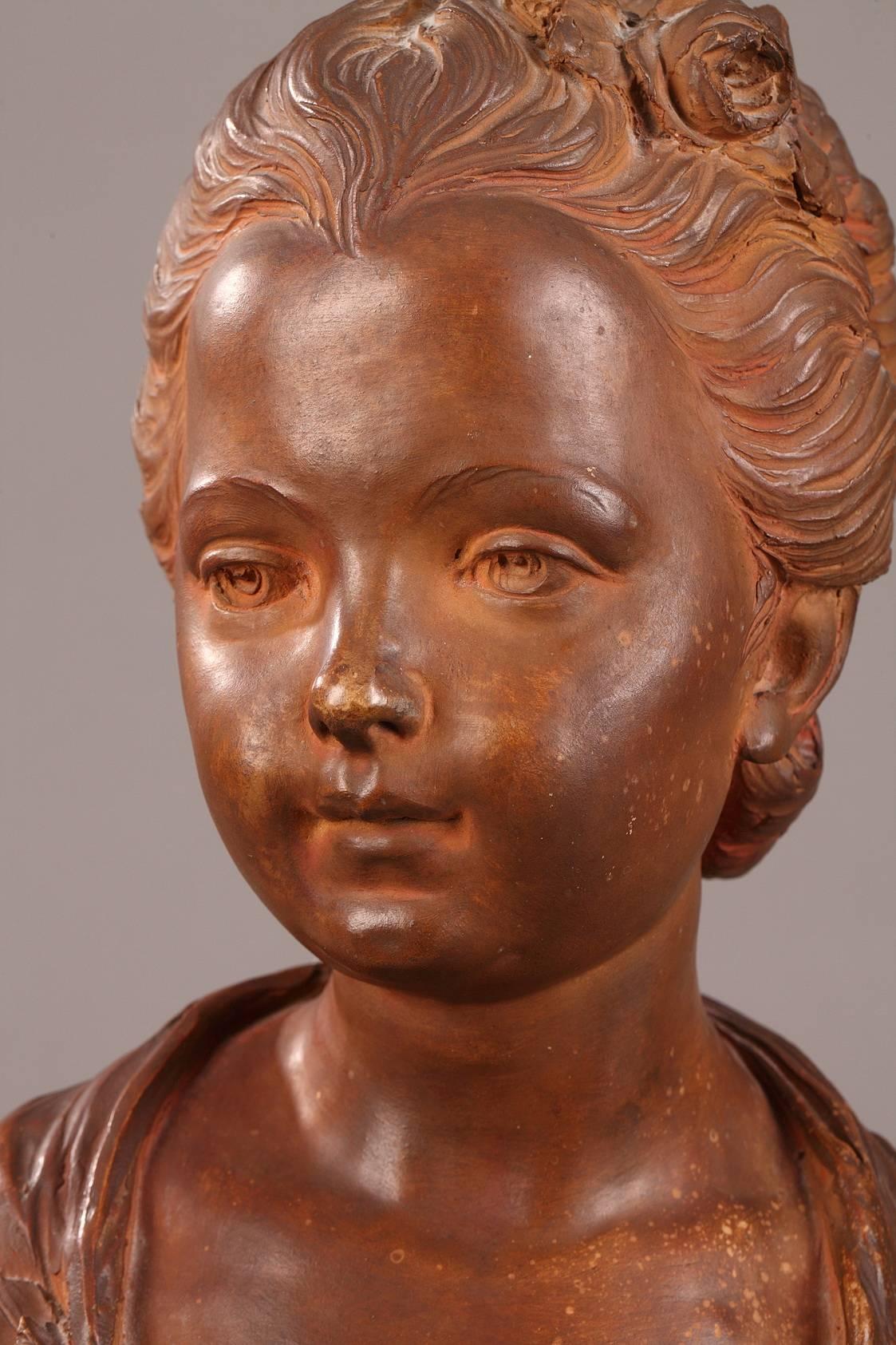 Terracotta bust of a young woman with a melancholy expression, dressed in 18th century clothing. Her hair, pulled back by a ribbon decorated with roses, frames a round and expressive face. The bust rests on a terraced, circular base. Signed on the