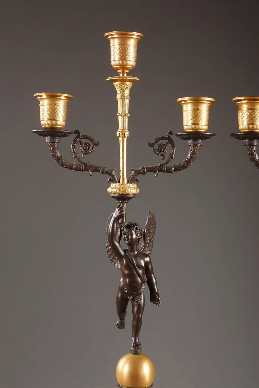 Pair of three-branched candelabras in gilded and patinated bronze. They each feature a winged, patinated bronze Cupid who is standing on a sphere and holding a torch with three candle branches. These patinated lateral arms are decorated with