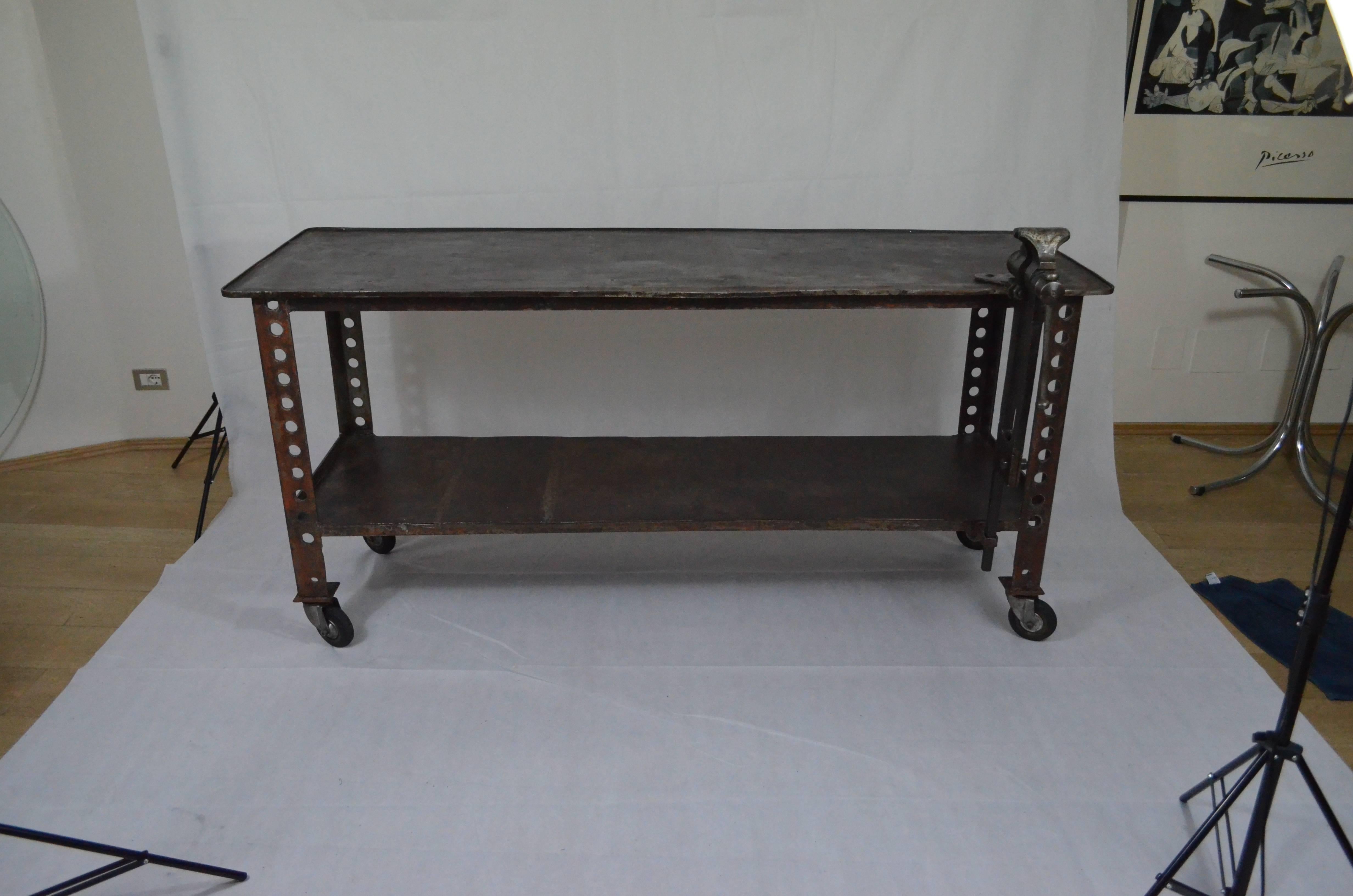 Iron Industrial work table form early 20th century from Italy. The pierced legs are directly connected with the top and on the right side there is a iron clamp perfectly working. There are two shelves, mounted on fours wheel, which end with an edge