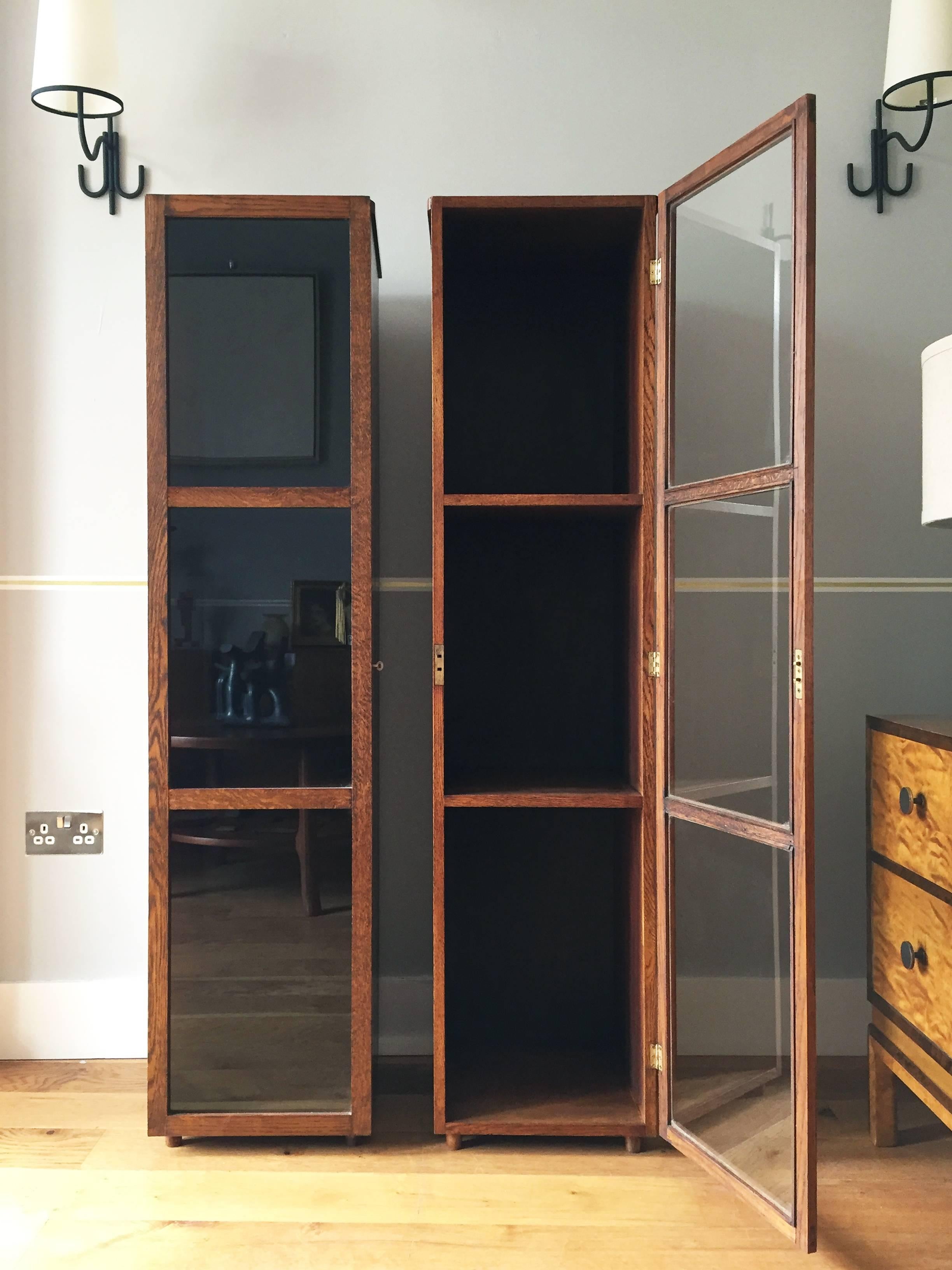 An unusual pair of oak narrow bookcases or display cabinets, divided in three compartments with shelves, closing symmetrically with glazed doors,
English, circa 1940.
Measures: 150.5 cm high by 36.5 cm wide by 35.5 cm deep.