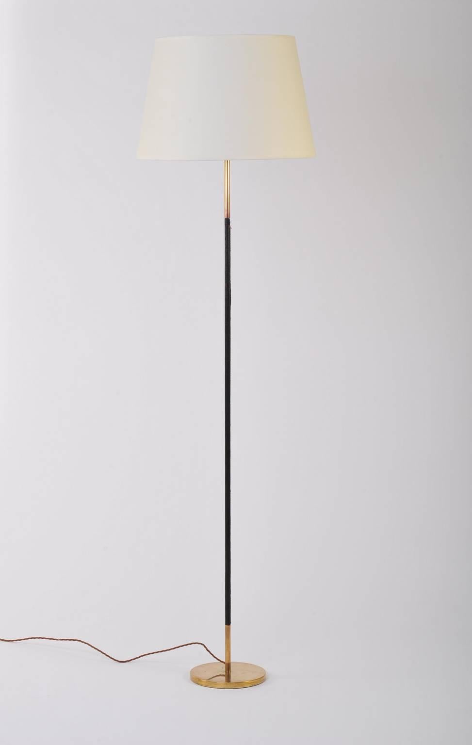 A brass and stitched black leather floor lamp, in the manner of Jacques Adnet, the elegantly thin stem supporting a bespoke ivory fabric shade
France, circa 1950.