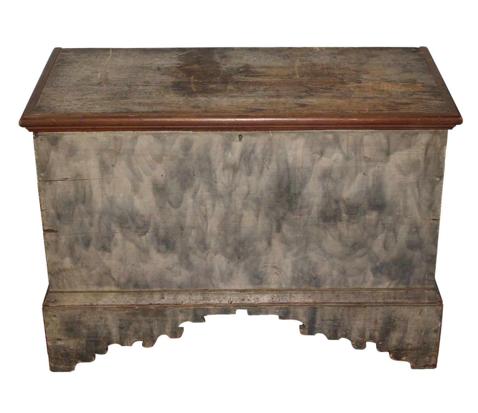 A beautifully smoke painted grey blanket chest, with decorative carving in front legs. On the inside, there are two smaller storage compartments, one cubby with a drawer underneath. There is a bit of space underneath the drawer as well. The hinges