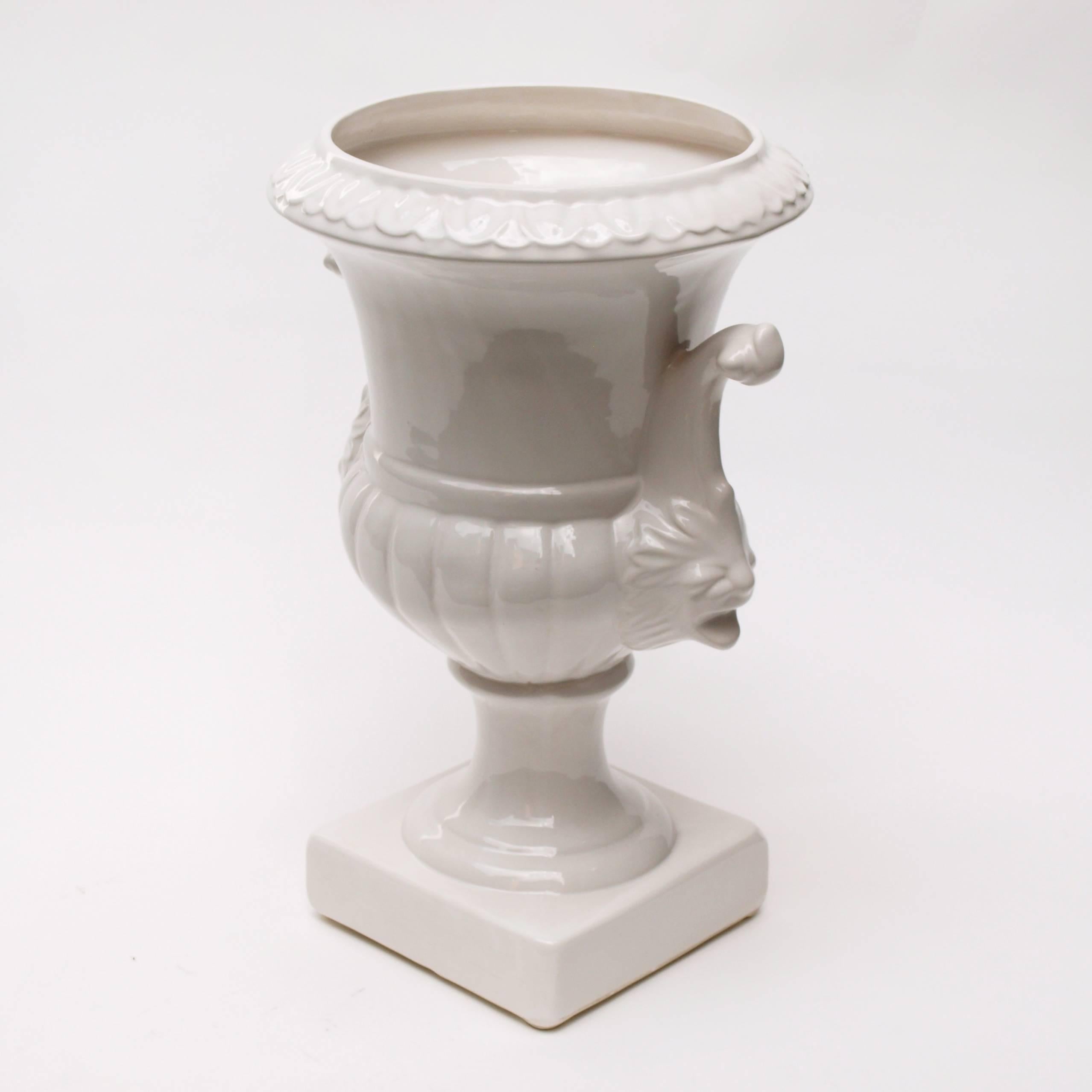 A large white glazed ceramic urn in a classical style, with lion head handle details and a leaf pattern circling the top of the vase. This sizable urn can be used outdoors or indoors as a planter or vase.