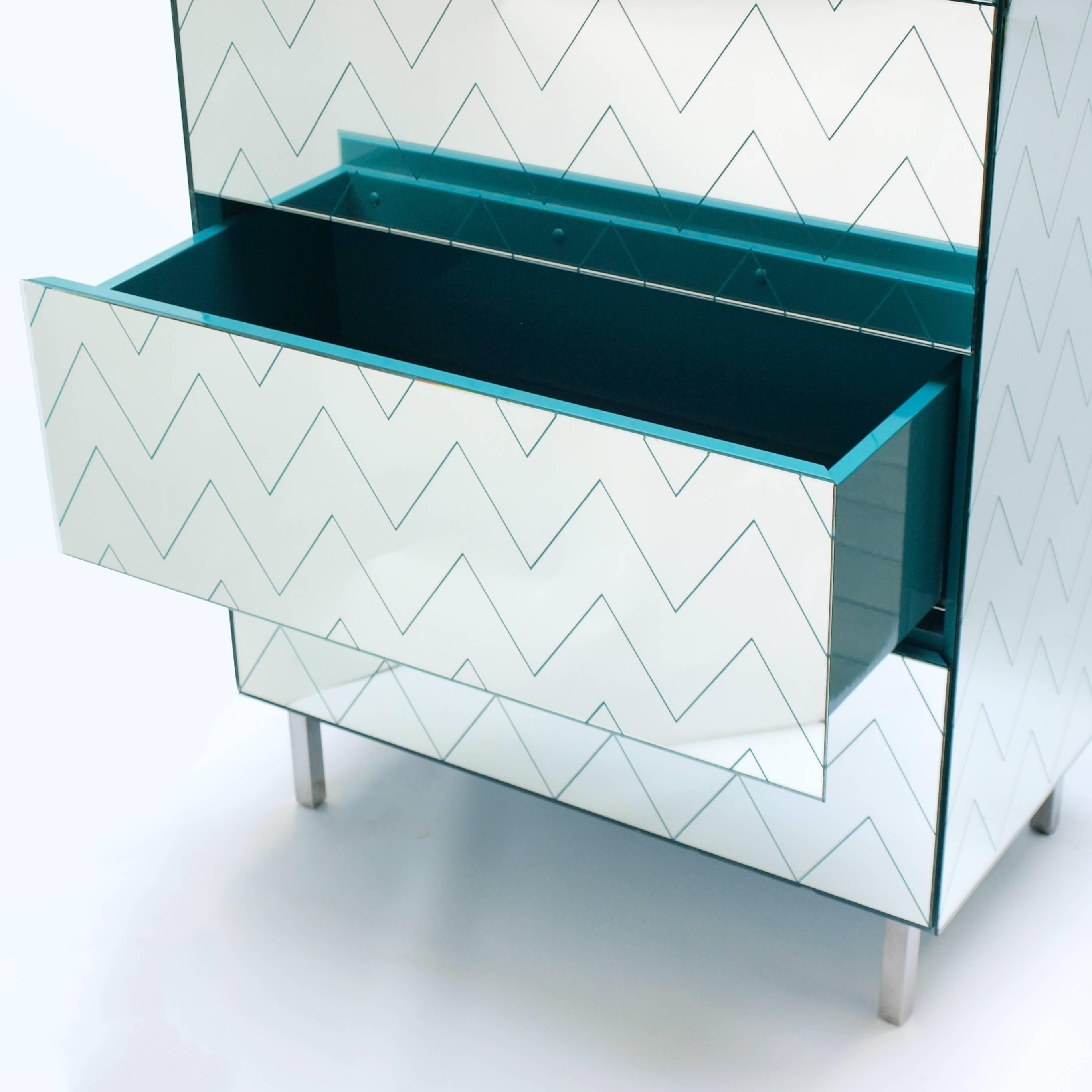 A fully mirrored glass chest of drawers adorned with turquoise coloured zig zags on four steel legs. Each drawer has a highly engineered soft close and opening mechanism and is finished inside with turquoise lacquer. The chest comes with a key