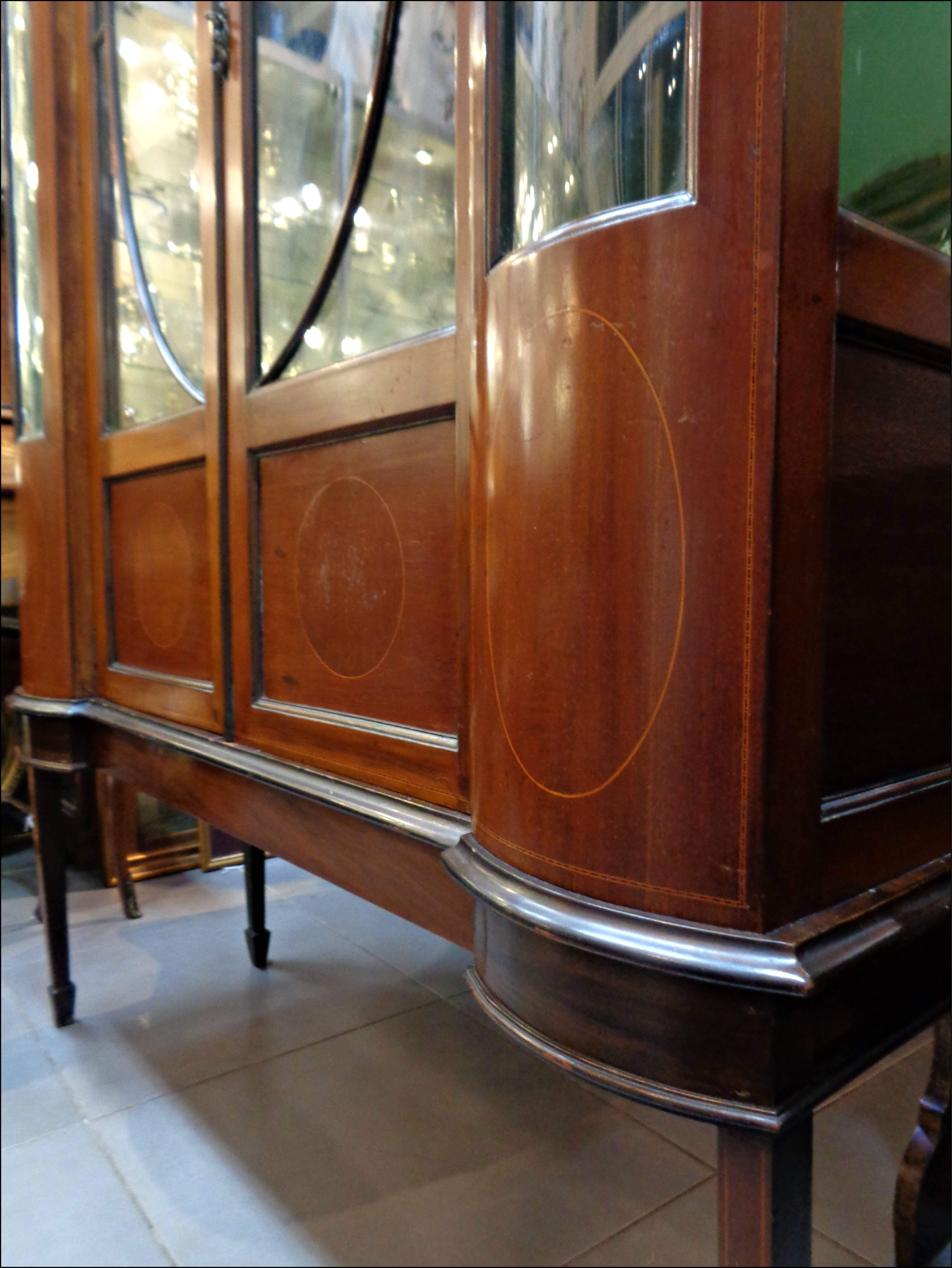 An English Edwardian glass display cabinet.
Made with fine mahogany veneer and classic frieze inlay 
with astragal glazed doors enclosing three shelves and tasteful bowed glass side wings, terminated with elegant tapering legs and a matching