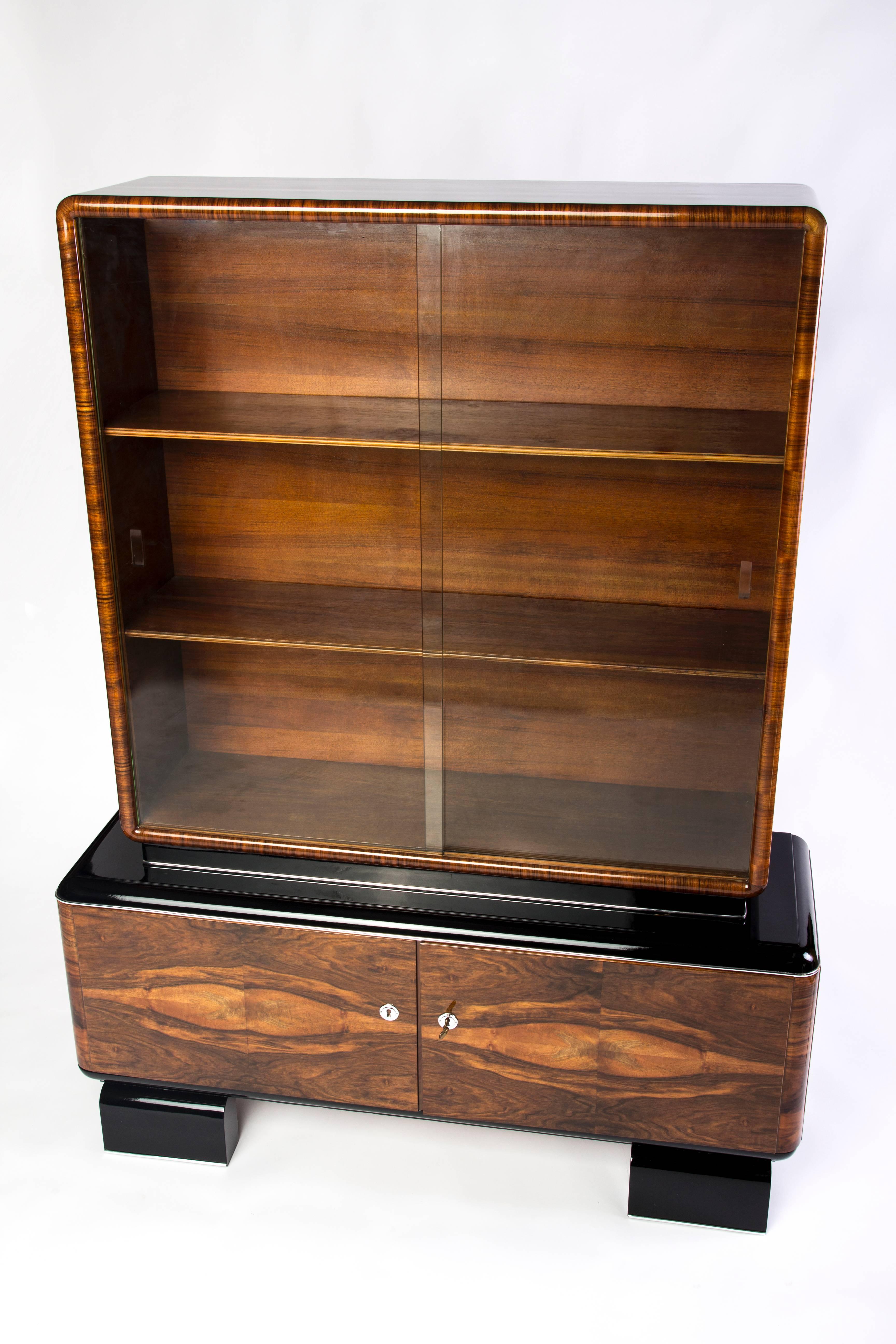 This magnificent Art Deco display case features hand polished burled wood, two (2) swing doors, and a high gloss black lacquer finished side panels and feet. The vitrine compartment features two (2) wooden shelves covered by a set of sliding