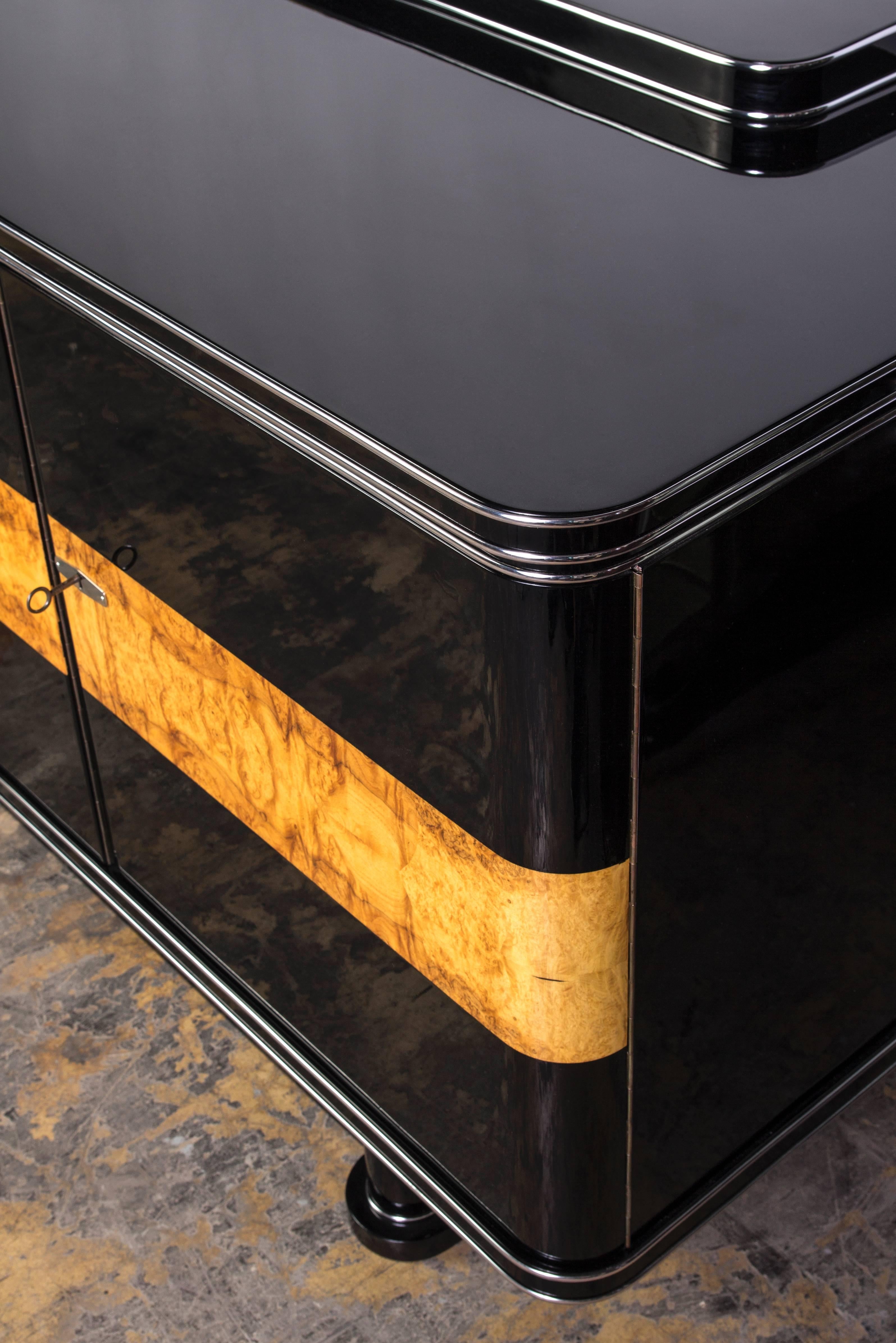 This colossal Art Deco sideboard depicts a very attractive two-tone combination of black piano lacquer and Loupe d’Amboine with chrome-line details. The interior is beautifully finished in cherrywood and burl for plenty of storage and drawers.