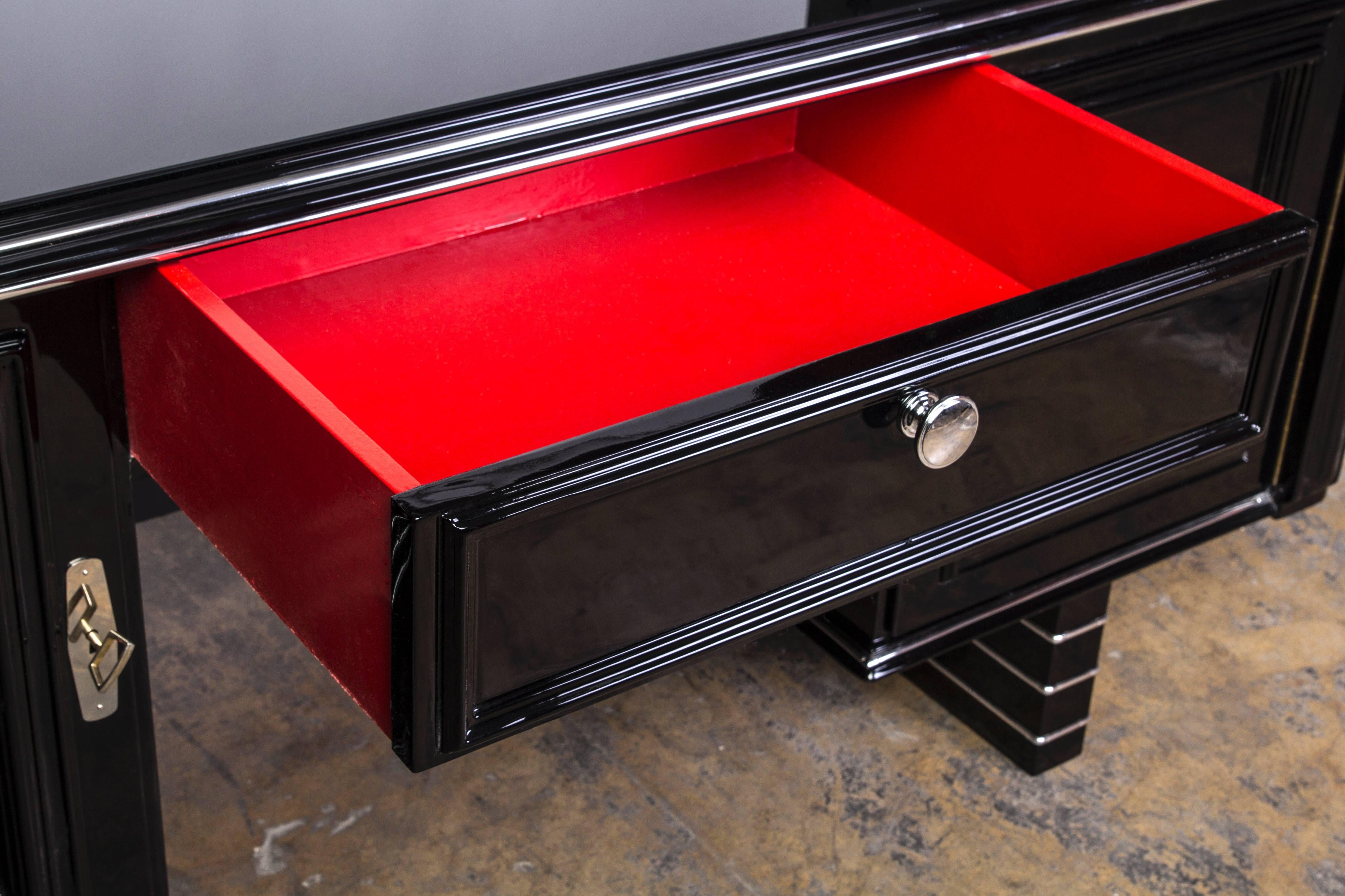 This magnificent Art Deco desk features two (2) swing doors with chrome lines and fixtures on a floating base design and a side to side shelve on the opposite side for display. Finished in a luscious high gloss black lacquer. Interiors in crimson