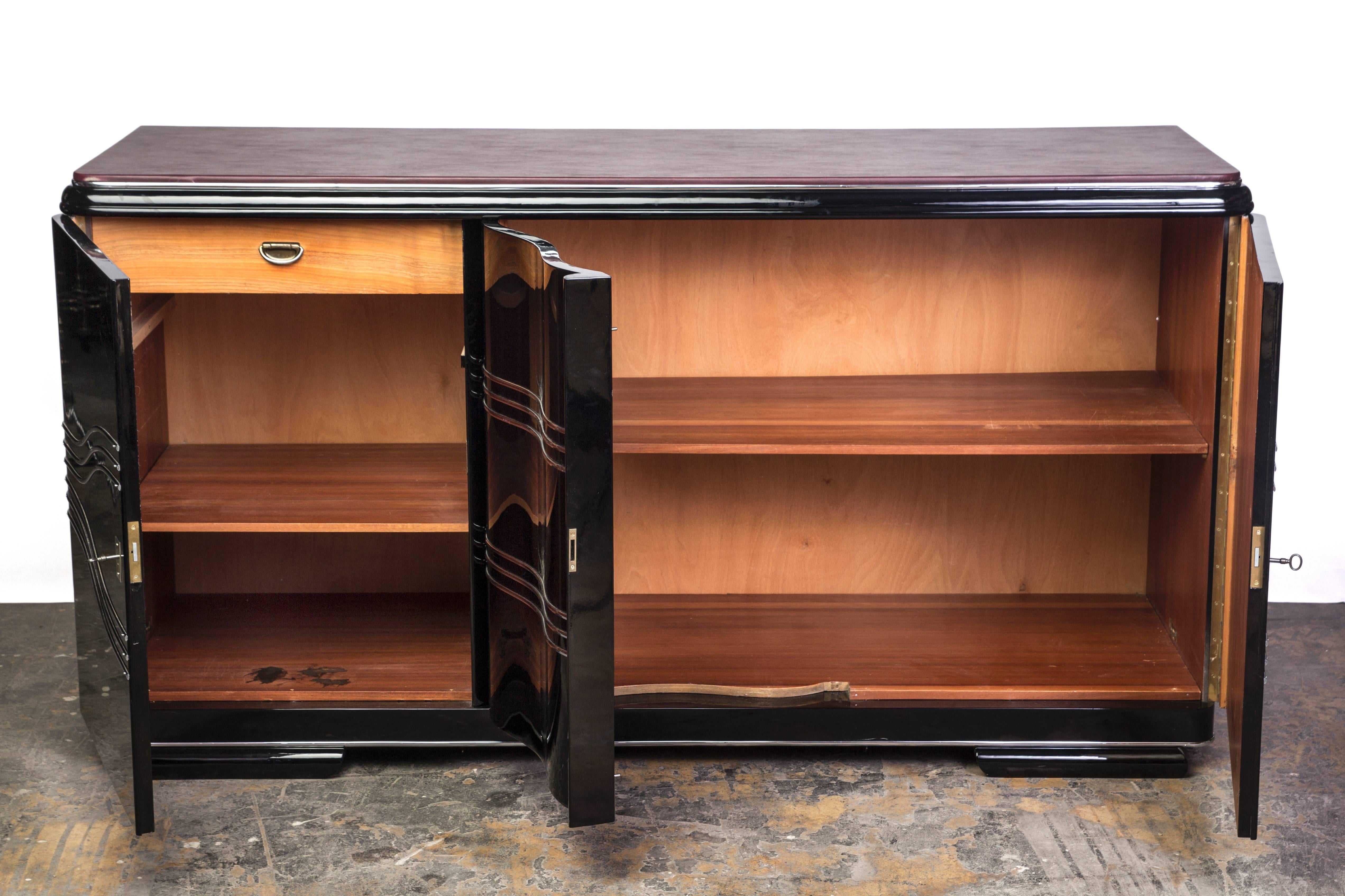 This gorgeous deco sideboard features three (3) swing doors with beautiful chrome detailing. It has a luscious leather top plate with a high gloss finish in black lacquer. Interiors mahogany.
   