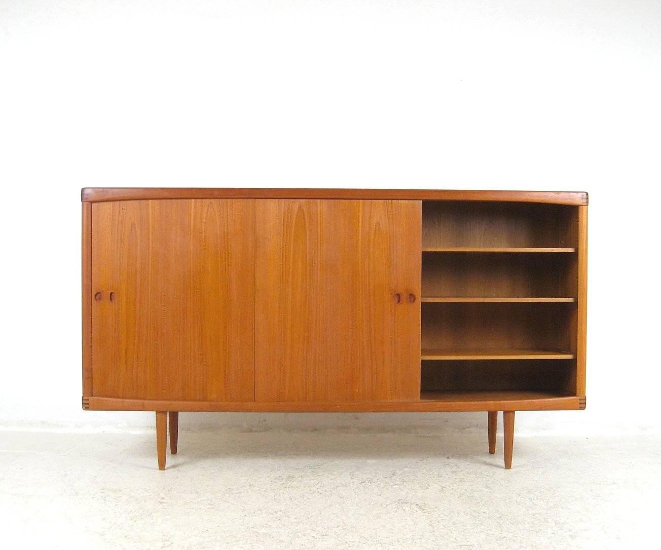 This teak sideboard was designed by H.W. Klein in the 1960s and manufactured by Bramin of Denmark. Made of teak heartwood, it comprises two cabinets behind a sliding door on each side and drawers in the centre. Above the drawers is a pull-down door