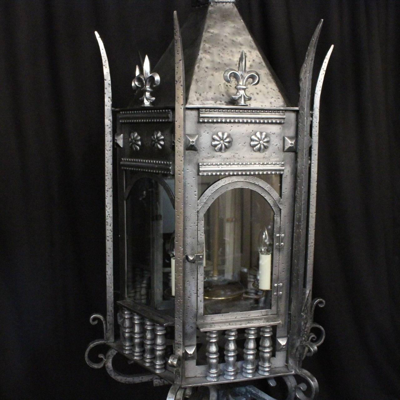 An English triple light wrought iron hexagonal Gothic antique lantern, the six glass panels held within a castellated framework with three internal light fittings, with square gauge spiked side sections, decorative floral embellishment and Fleur de